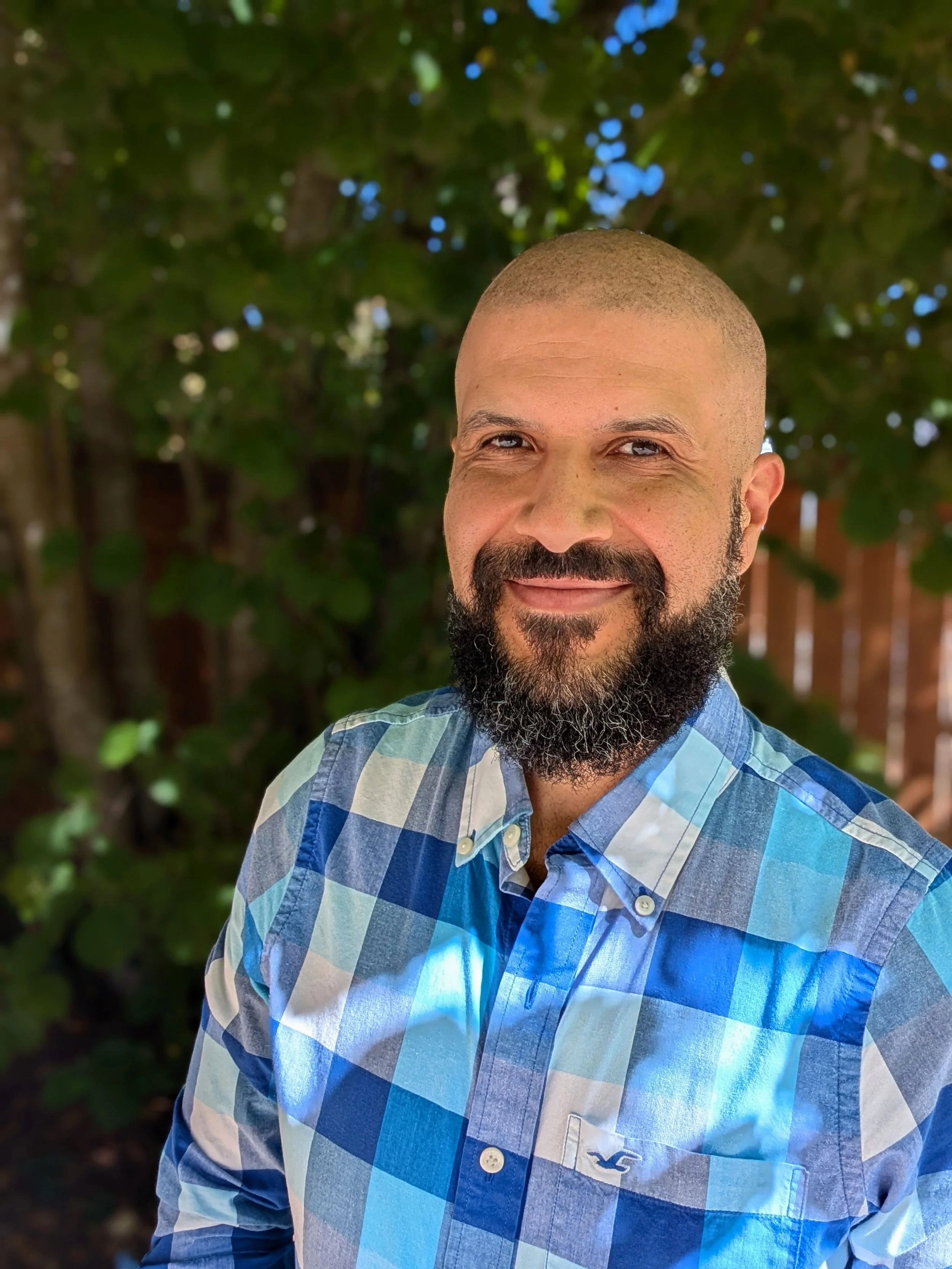Dr. Correia with a beard and a bald head smiling outdoors, wearing a blue and white plaid shirt, with a blurred green leafy background.