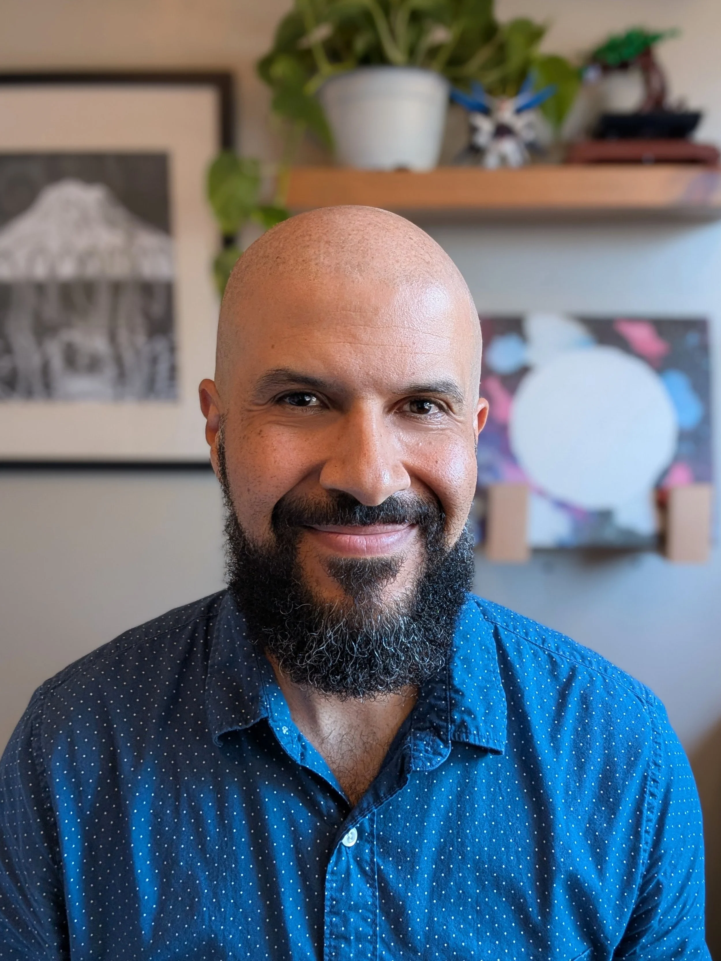 Close-up of Dr. Correia with a shaved head and beard, wearing a blue shirt with white polka dots, in a room with framed pictures, vinyl records, and a plant in the background.