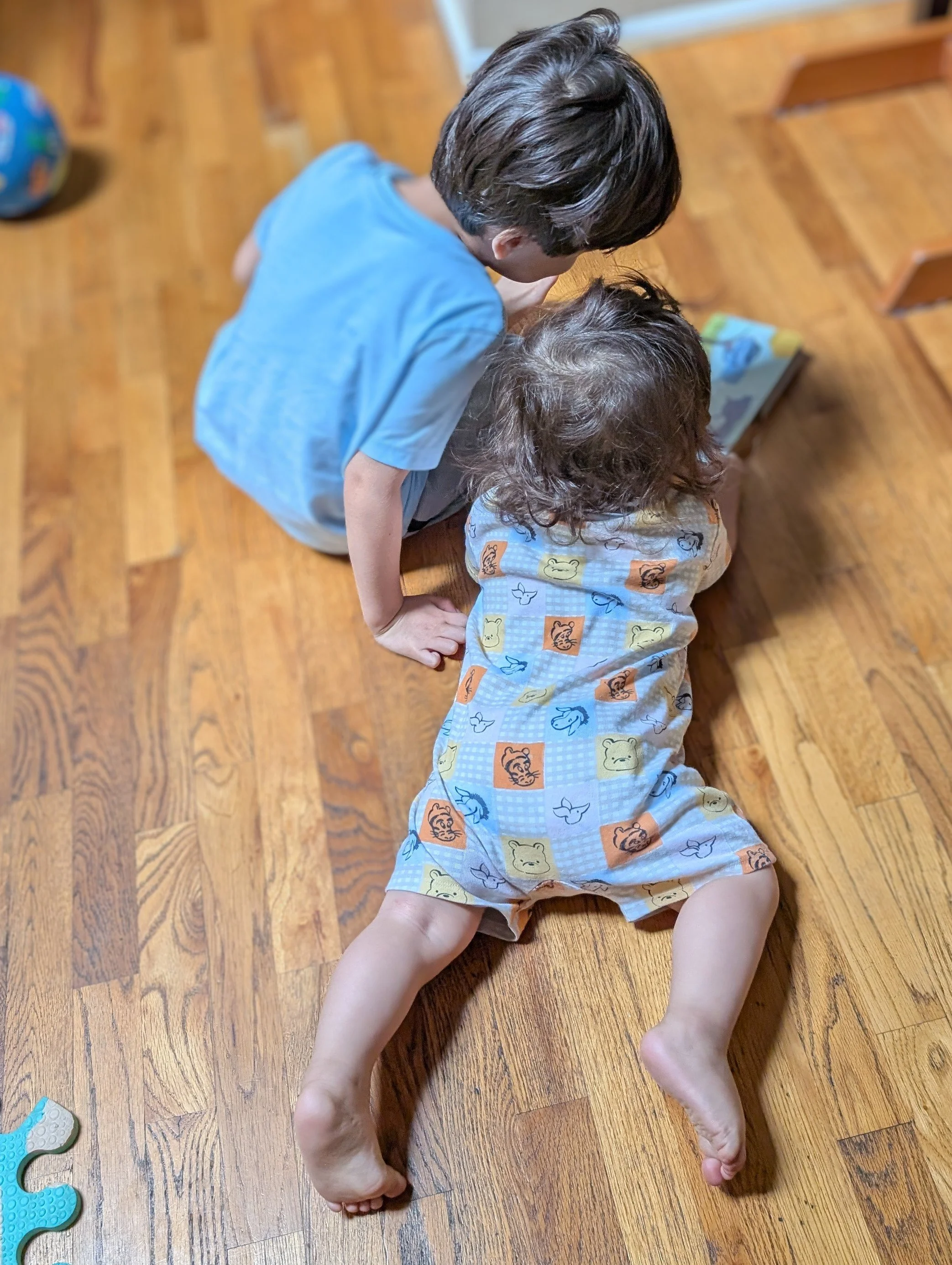 Dr. Correia's two children are playing on a hardwood floor, one is lying on the floor wearing a colorful animal-printed shirt and the other is kneeling over them wearing a blue shirt.