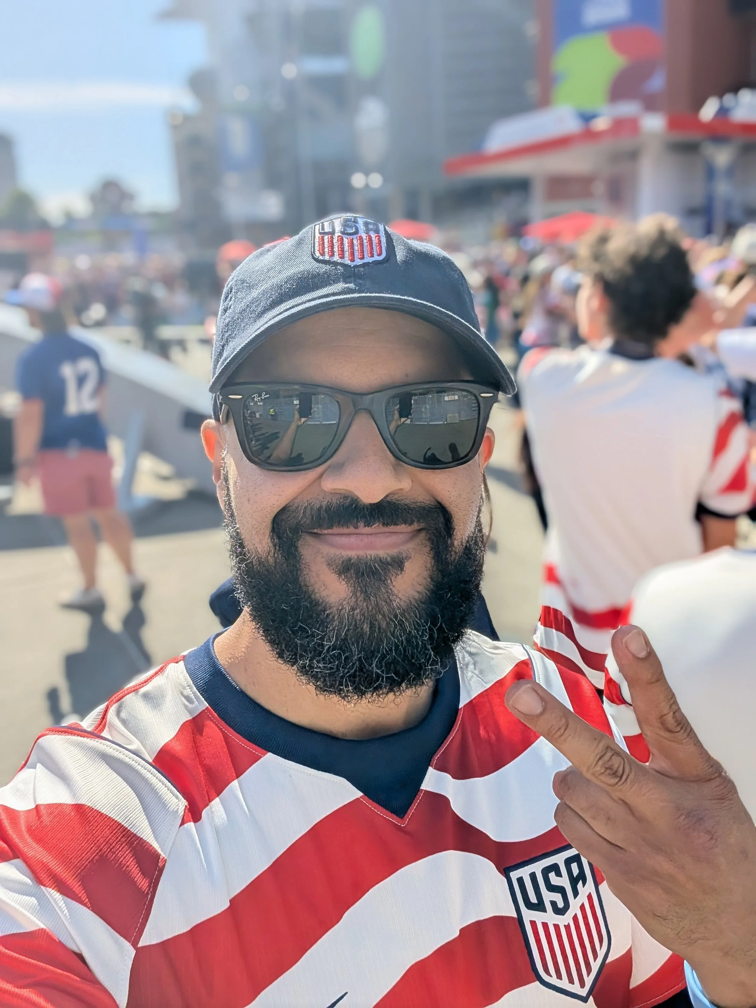 Dr. Correia wearing sunglasses and a US soccer jersey is smiling and making a peace sign with his right hand at World Cup game with a crowd in the background.