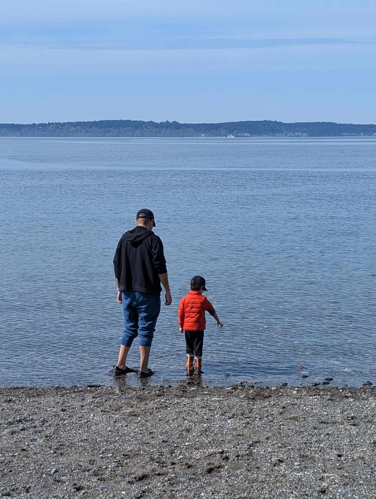Dr. Correia and his son walk along the foreshore of the Puget Sound on a cloudy day, with Dr. Correia wearing a black jacket and cap, and his son wearing a red jacket and colorful rain boots, both with their feet in the water.