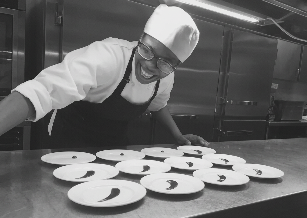 A chef in a white uniform and hat, smiling broadly, stands behind a counter with multiple plates, each holding a curved, dark edible item, in a professional kitchen with stainless steel appliances.