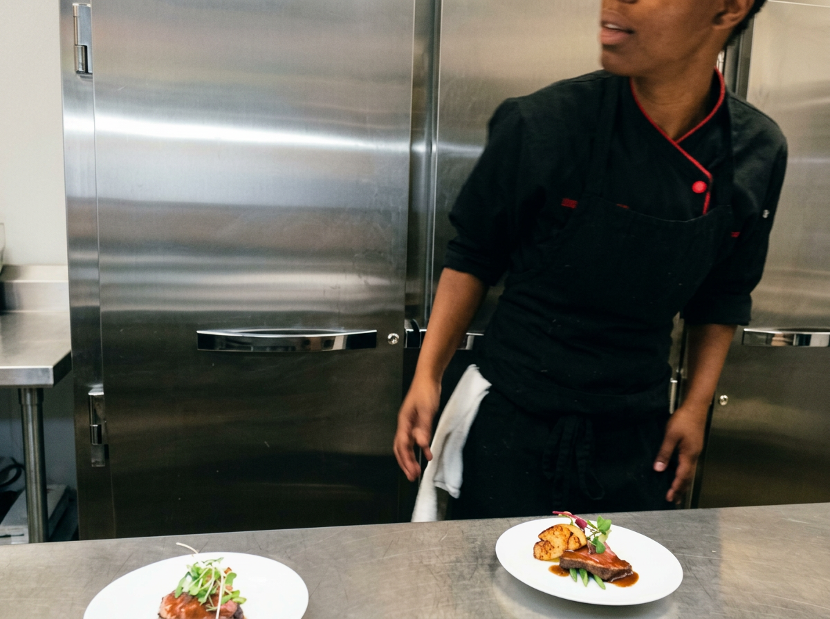 Chef in black uniform stands behind stainless steel counter with two plated dishes of food, including meat, vegetables, and garnishes.