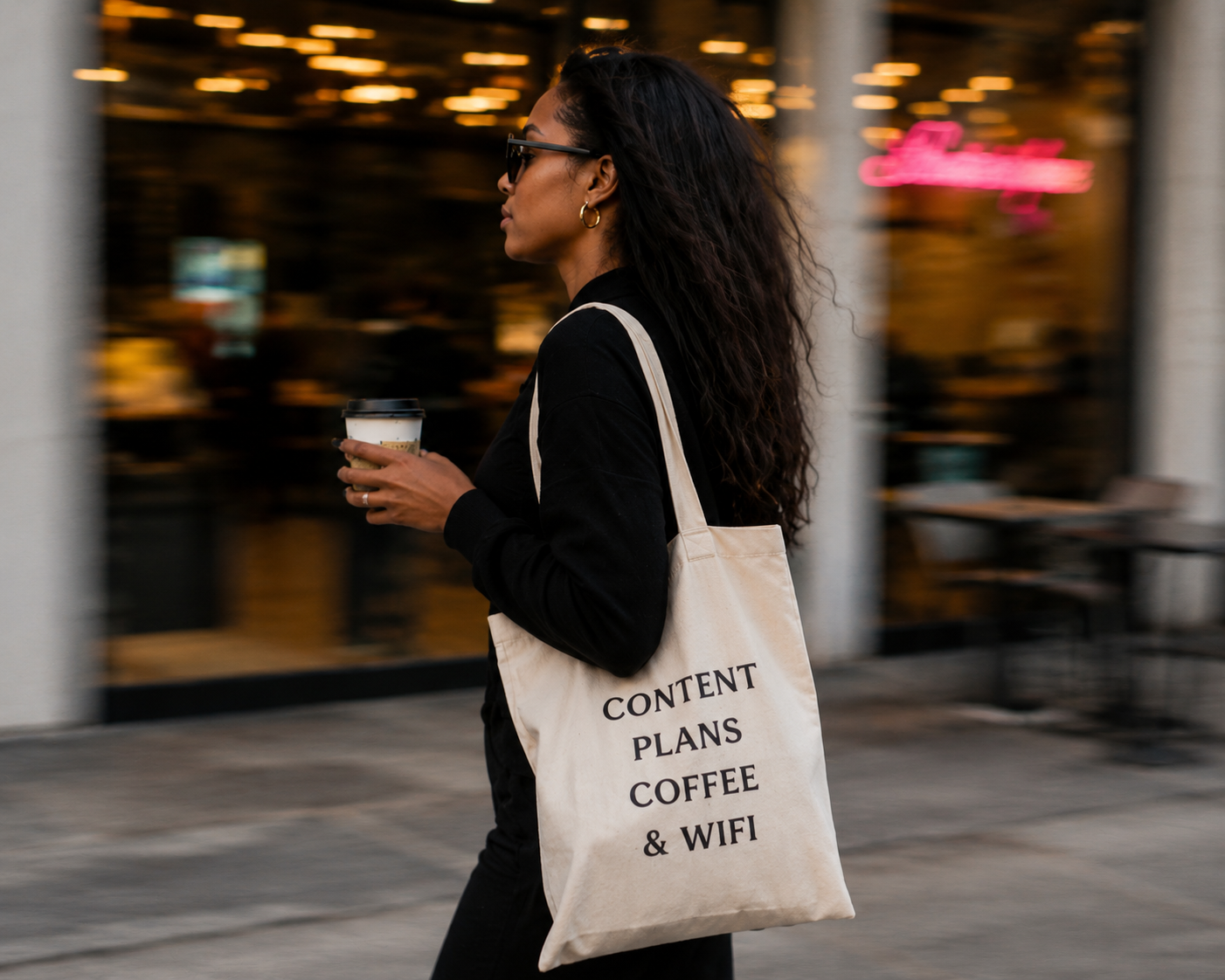 A woman with curly hair and glasses walking outside at dusk, carrying a coffee cup and a tote bag that reads 'Content Plans Coffee & WiFi.'
