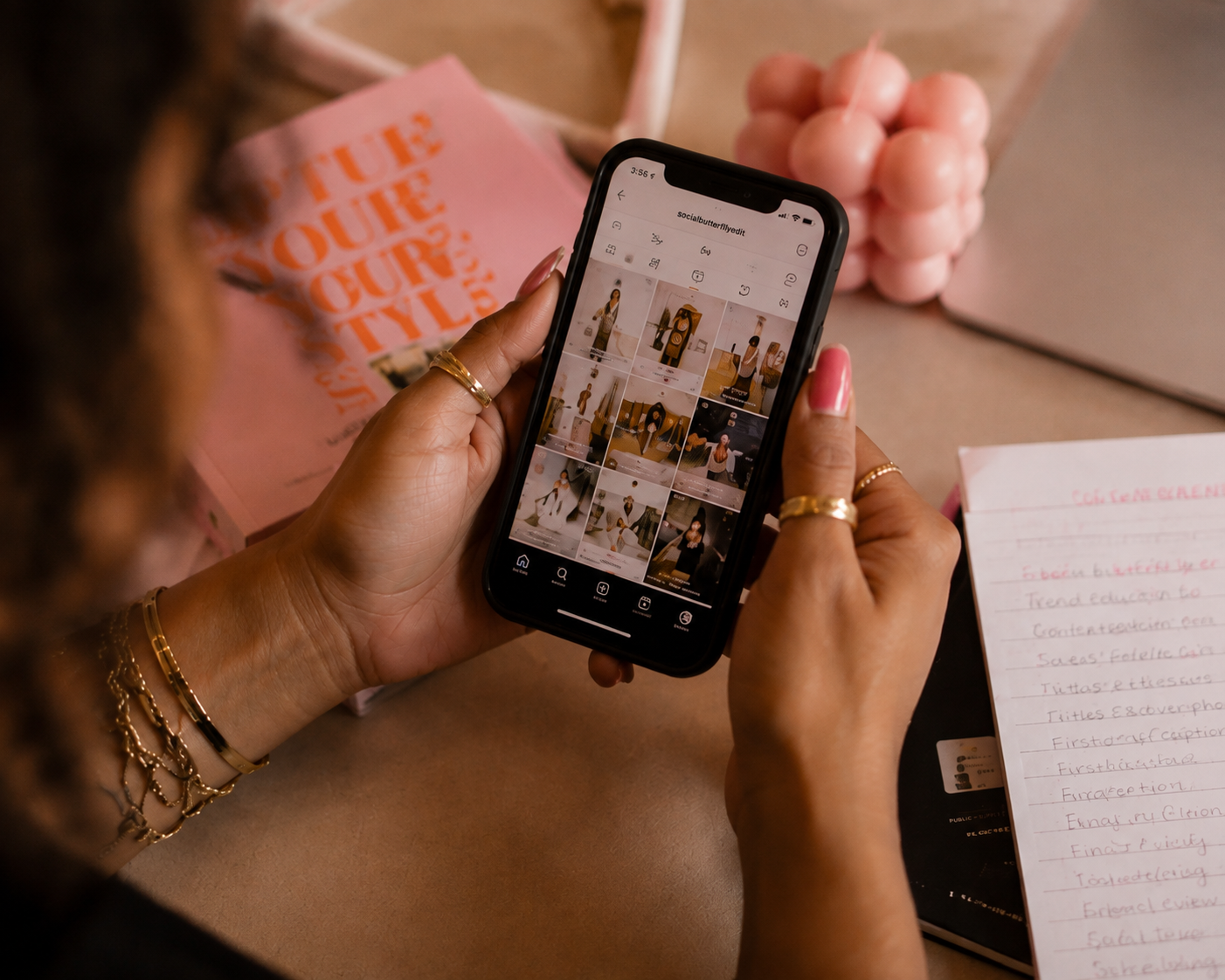 Person viewing a grid of fashion images on a smartphone. There are pink notebooks and a notepad with handwritten notes nearby.