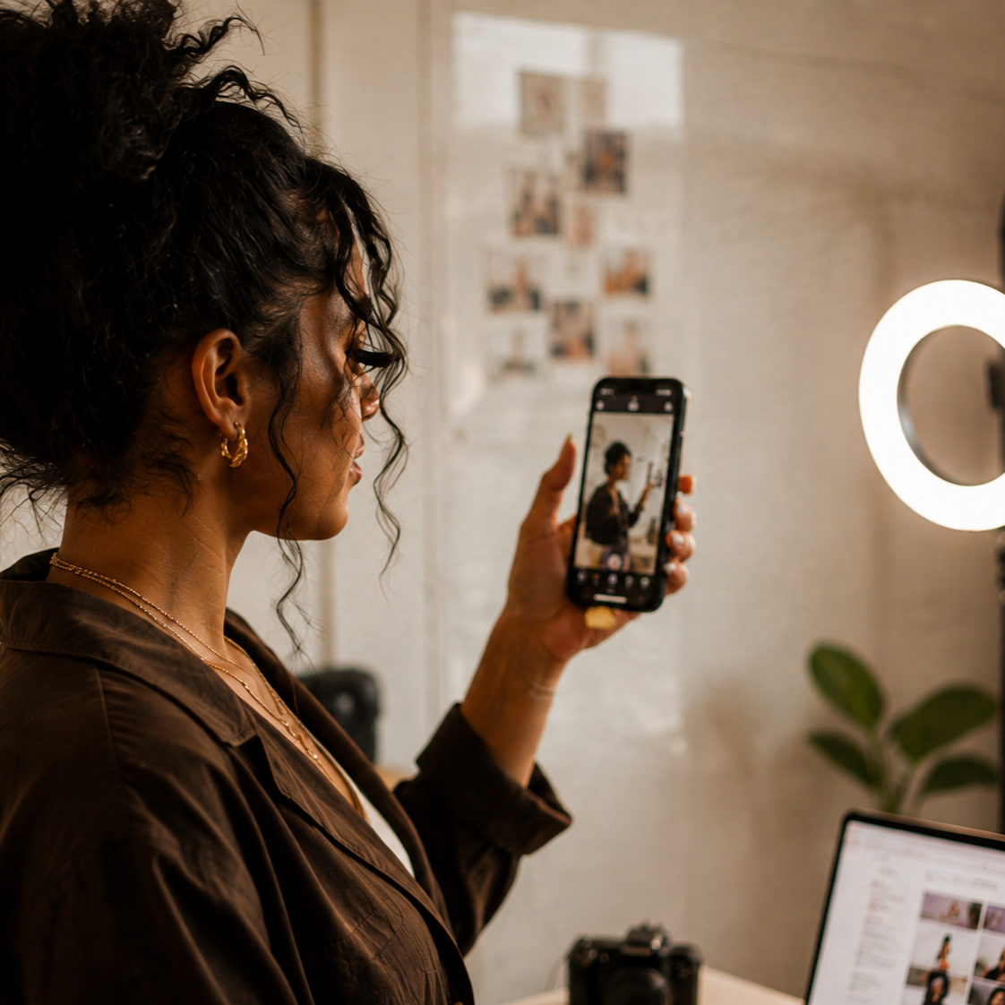 Content creator filming with a smartphone beside a ring light, laptop, and camera in a creative workspace.