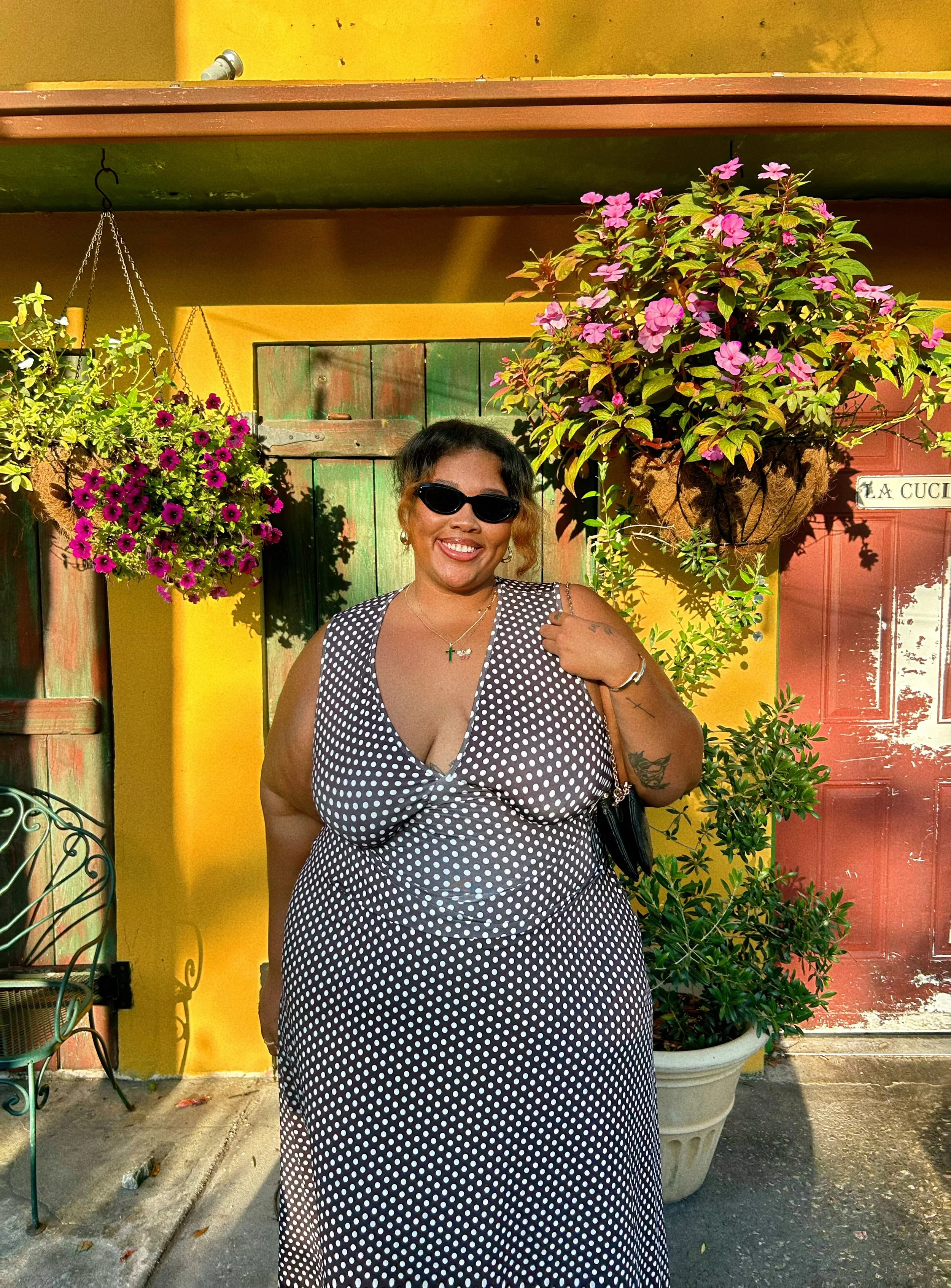 A smiling woman with dark sunglasses, wearing a black and white polka dot dress, standing in front of a yellow wall with potted pink and purple flowers, some on hanging baskets.