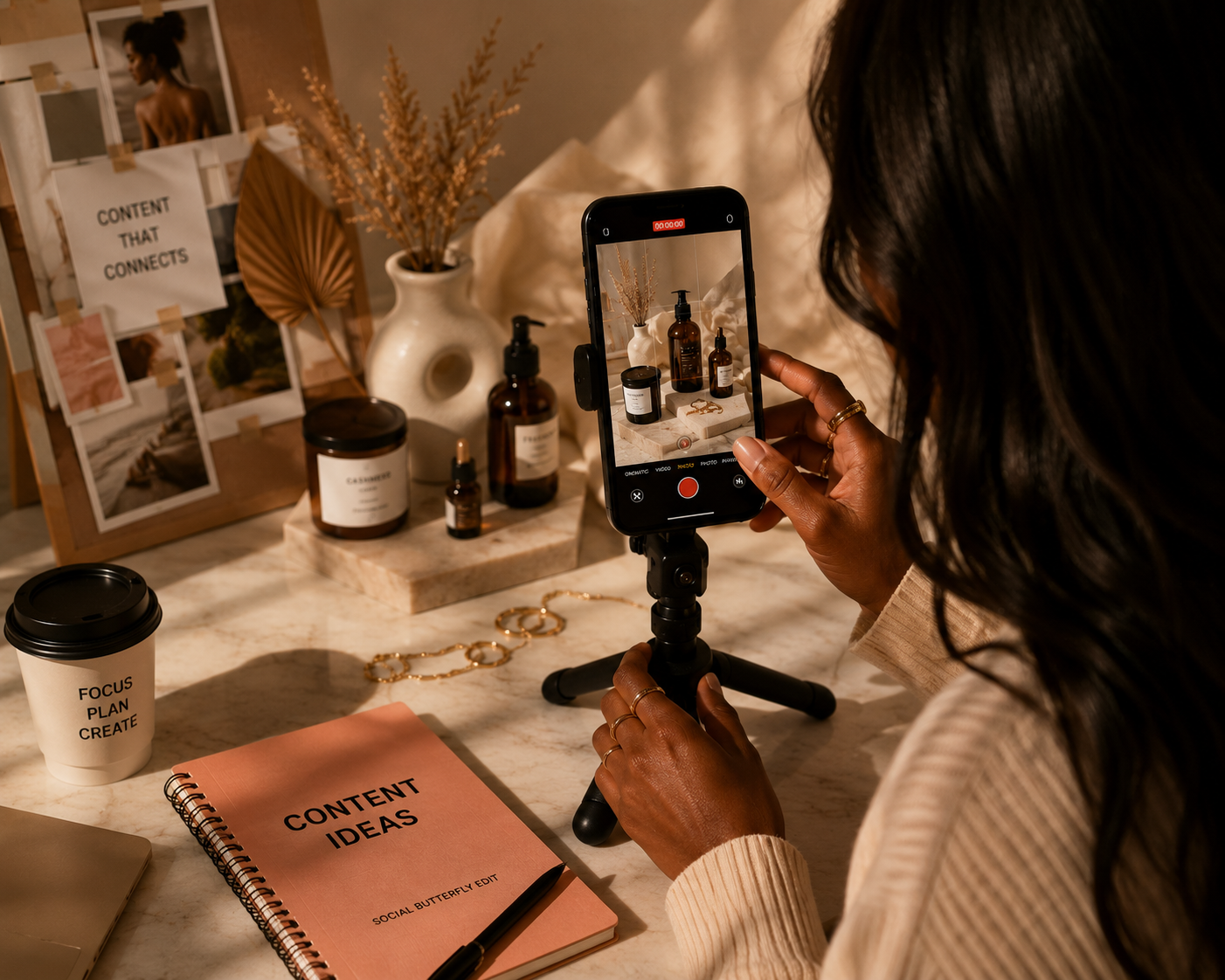 A person filming a table with various cosmetic bottles and a notebook labeled 'Content Ideas' using a smartphone on a tripod. The table also has a coffee cup labeled 'Focus Plan Create' and a jewelry piece, with a decorative board of photos and text in the background.