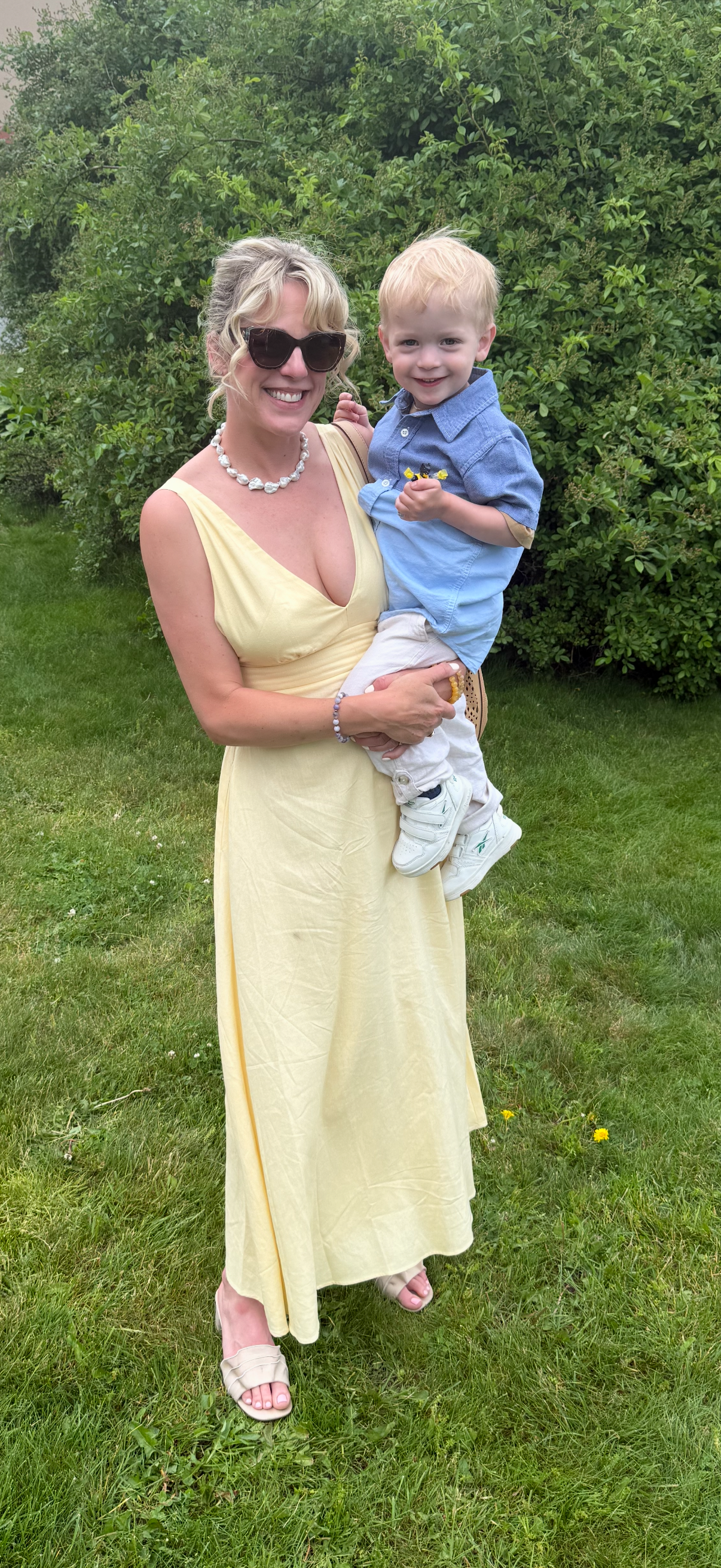 A woman wearing beige dress, sunglasses, and jewelry holding a smiling young boy in a blue shirt and white pants outdoors near green shrubbery.