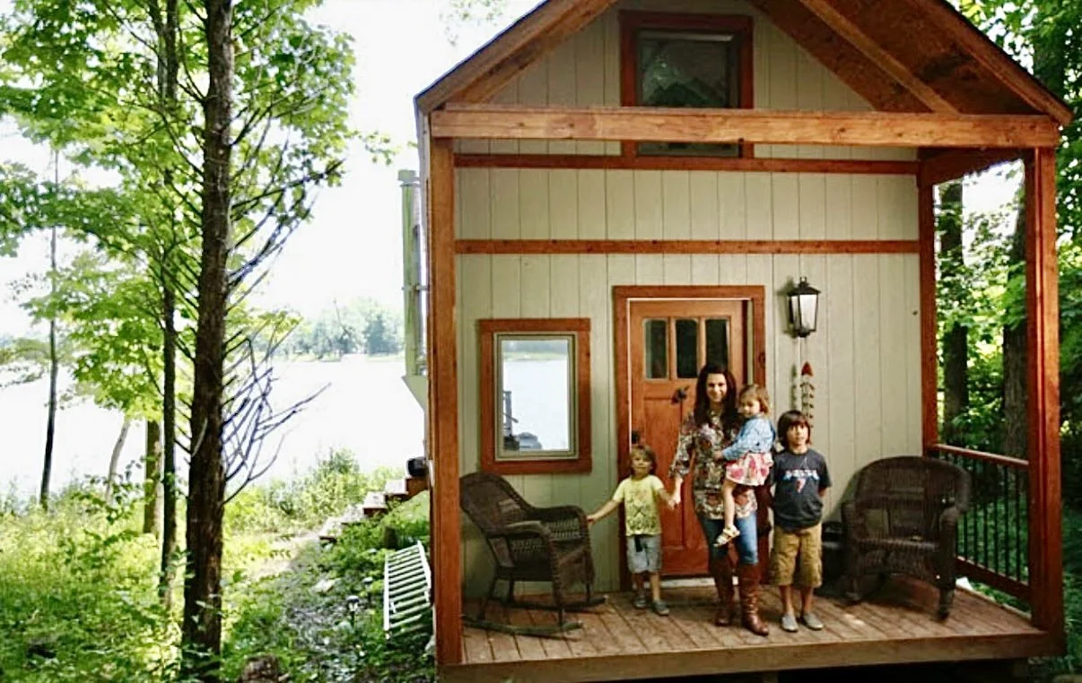 A small woman holding a young girl on a porch with three children, two boys and a girl, standing next to her in front of a house near a wooded lake.