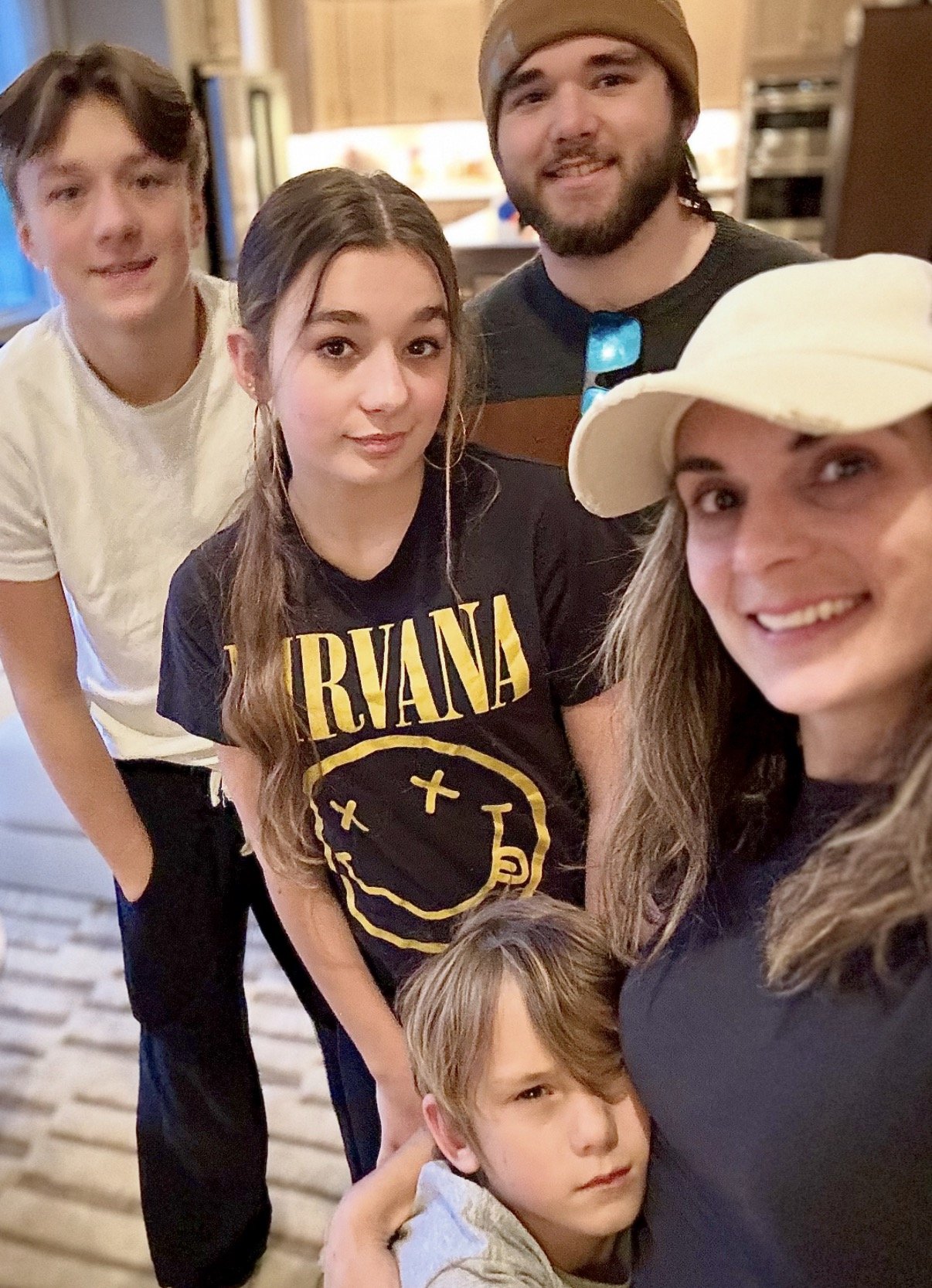 A group of five people, three women, one man, and one boy, smiling and taking a selfie together indoors. The woman in the front right is wearing a white cap, and the woman in the middle has long wavy hair and is wearing a black T-shirt with a Nirvana logo. The man behind her is wearing a beanie. The boy in front has light brown hair and is looking at the camera.