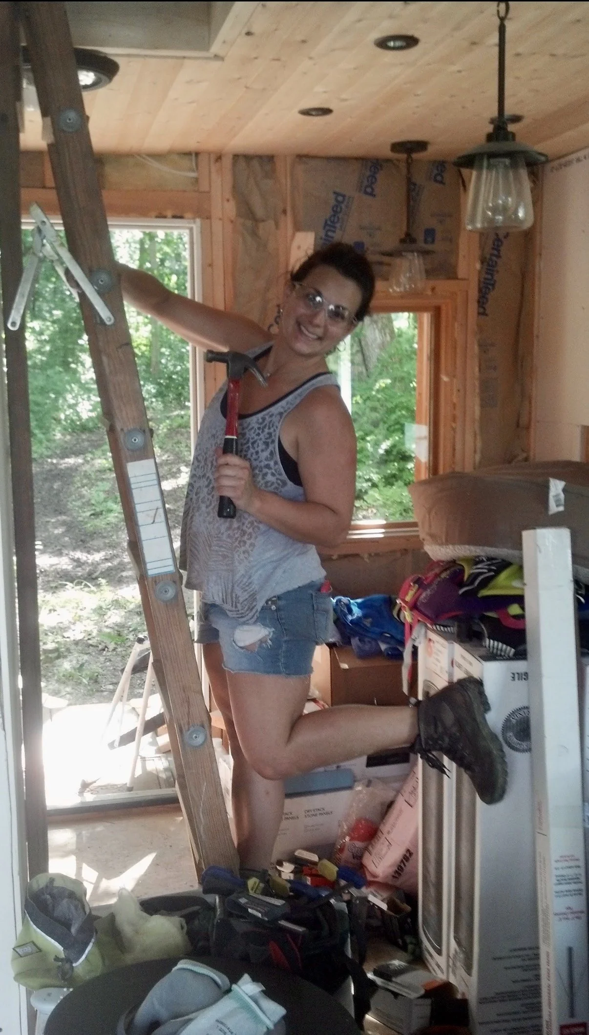 A woman with short dark hair, glasses, a sleeveless patterned top, and ripped denim shorts, standing on a ladder inside a room under construction. She is holding a hammer, smiling, with one leg raised and foot resting on a nearby object. Tools and construction supplies are scattered around.