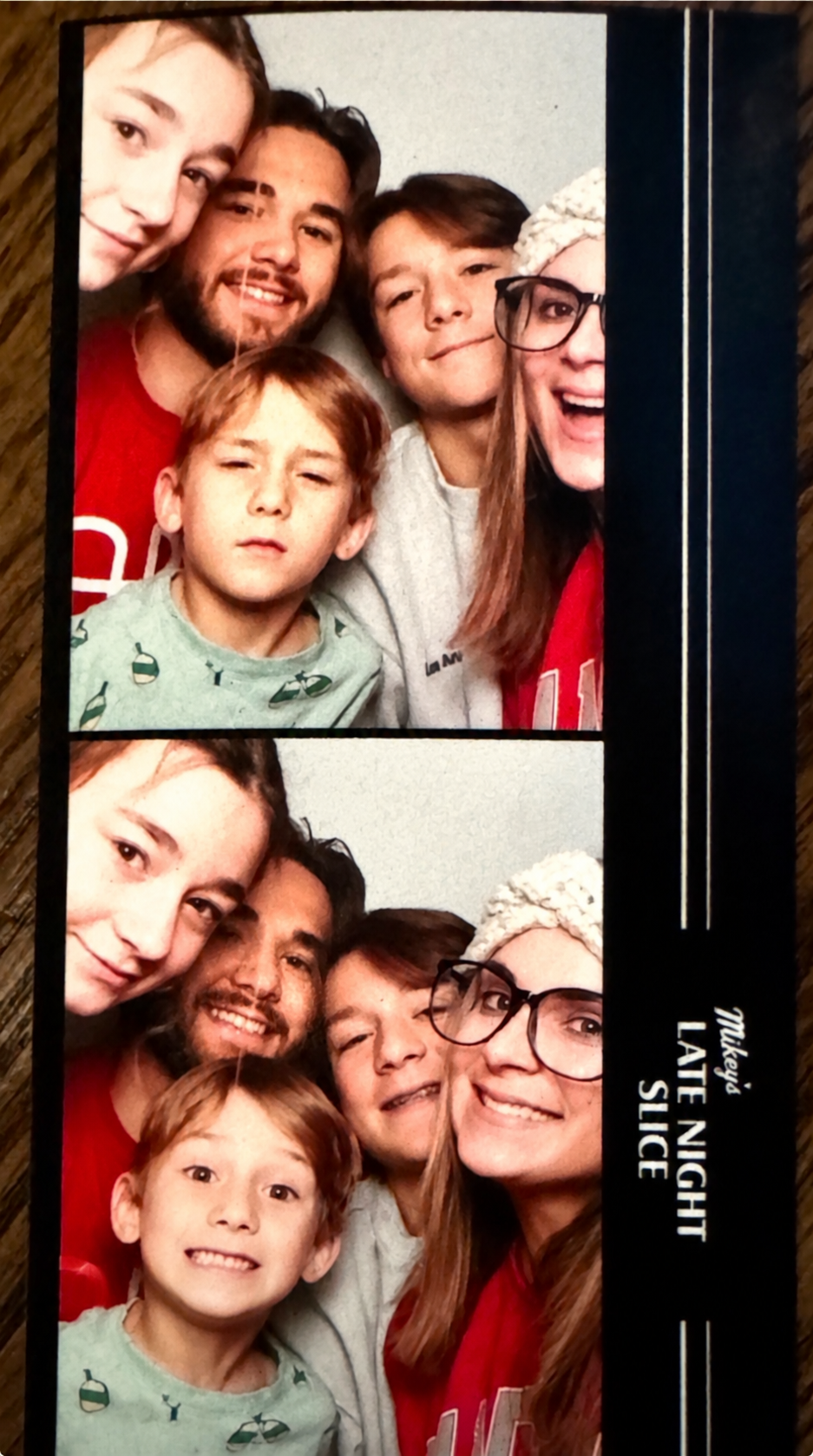 Family taking a group photo in a photo booth, with two pictures stacked vertically. The family includes a woman with glasses and a headband, two boys, a man with a beard, and a young boy with red hair. The top photo shows most of them smiling and making different expressions, while the bottom photo has some expressions changed.