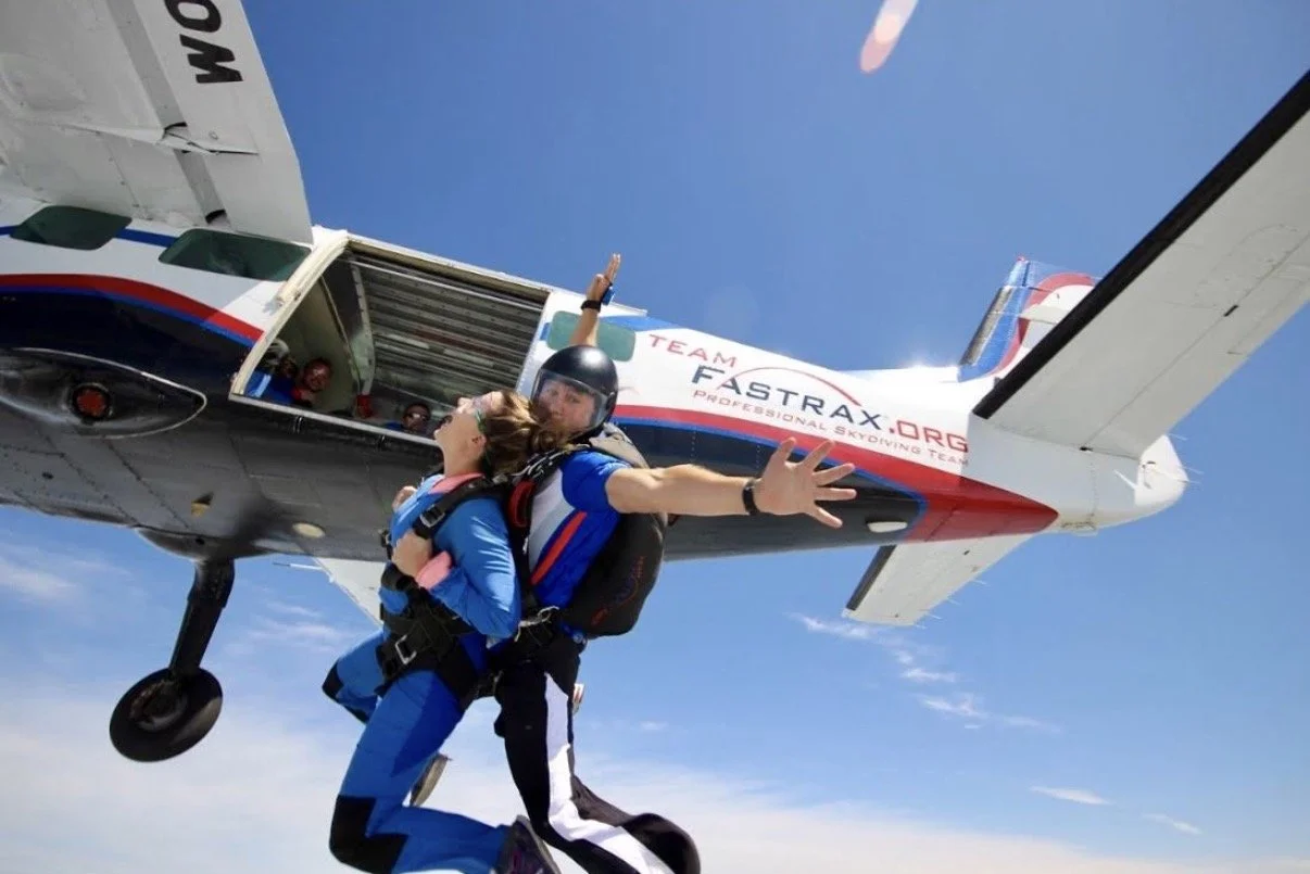 Skydivers exiting an airplane in mid-air, with one skydiver reaching out towards the camera, under a clear blue sky.