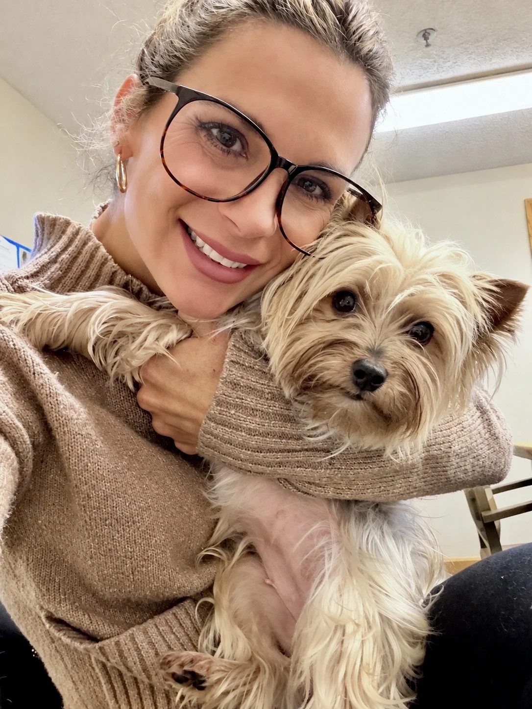 A woman with glasses smiling and holding a small, light-colored dog with long, slightly curly fur, indoors.