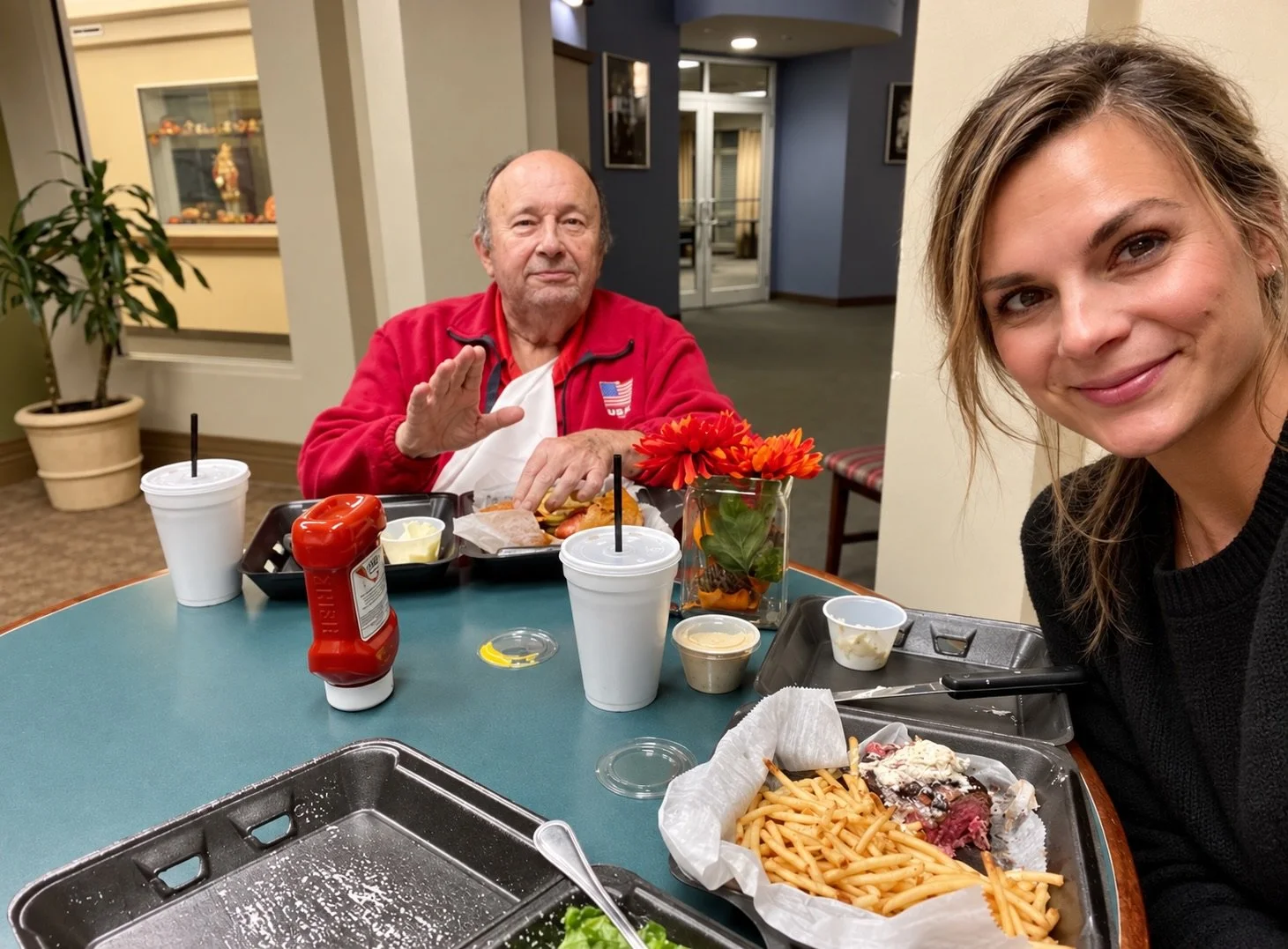 A man and a woman sit at a table in a cafeteria, with food trays, drinks, and condiments. The man is wearing a red jacket with a U.S. flag patch and is gesturing with his hand, while the woman is smiling at the camera with spaghetti and a salad in front of her.