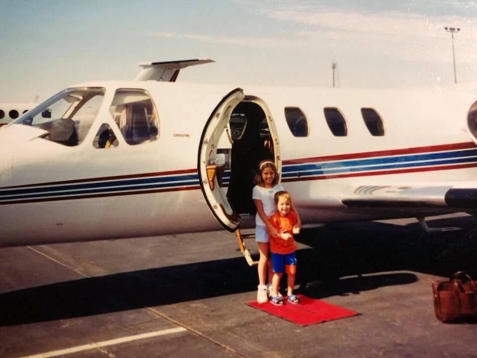 Jenny Ann Urban as a child with her brother standing in front of a private aircraft, reflecting a lifelong connection to aviation.