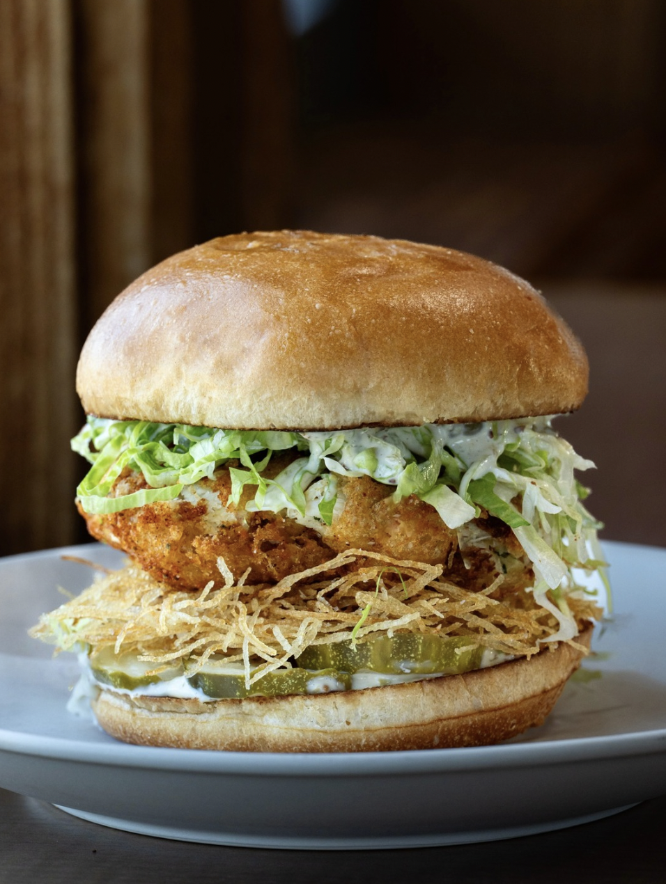 Close-up of a fried fish sandwich with lettuce, pickles, crispy onions, and sauce in a sandwich bun on a white plate.