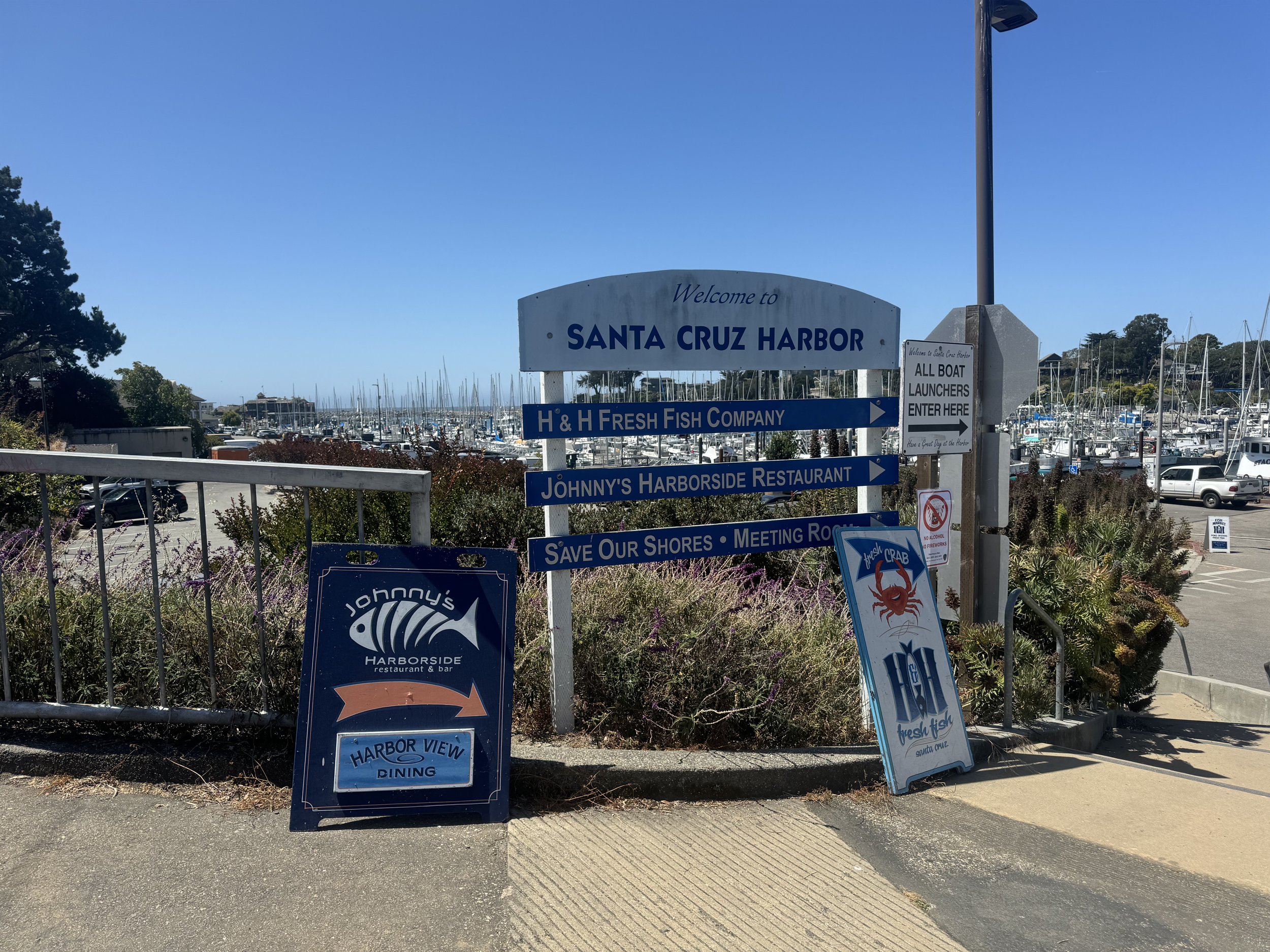 Welcome sign for Santa Cruz Harbor with directional signs for H & H Fresh Fish Company, Johnny's Harborside Restaurant, and Save Our Shores Meeting Room, with boats docked in the harbor and a parking lot in the background.