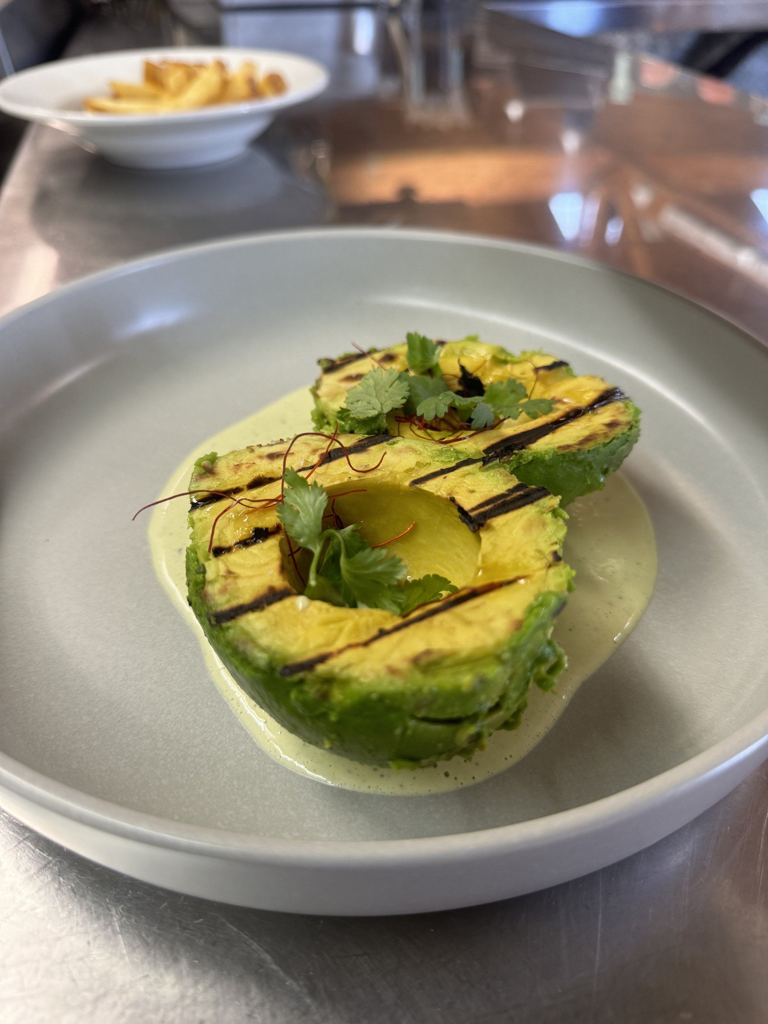 Grilled avocado halves garnished with cilantro and red chili threads in a light green sauce on a white plate.