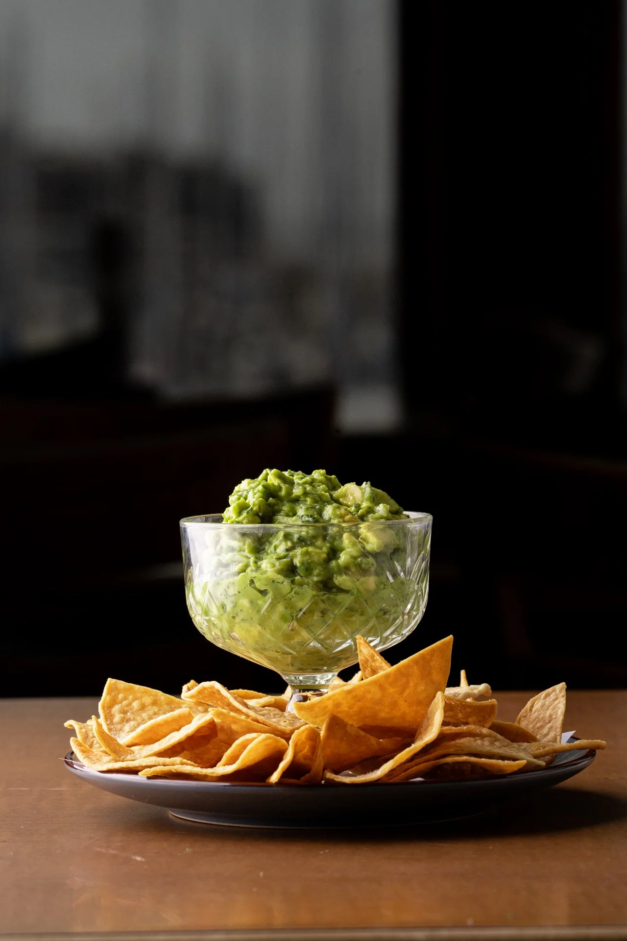 A glass bowl filled with guacamole placed on a black plate surrounded by fried tortilla chips, on a wooden table with a dark blurred background.