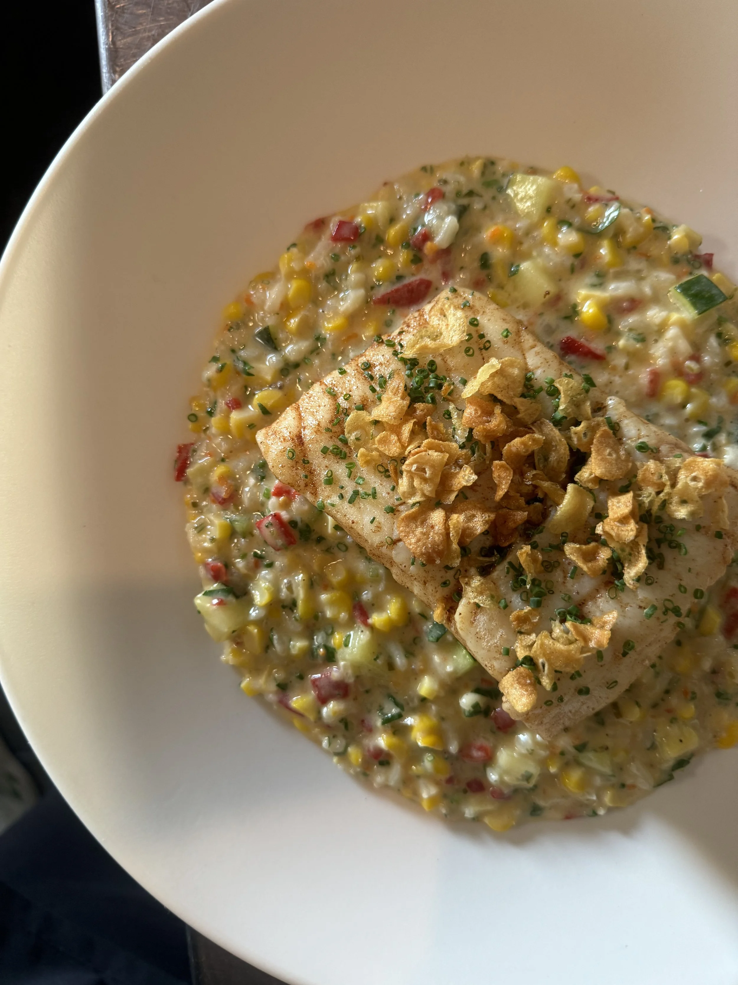 A white bowl containing a fish fillet topped with crispy garlic bits, served over a bed of creamed corn with red bell peppers, corn, and green herbs.