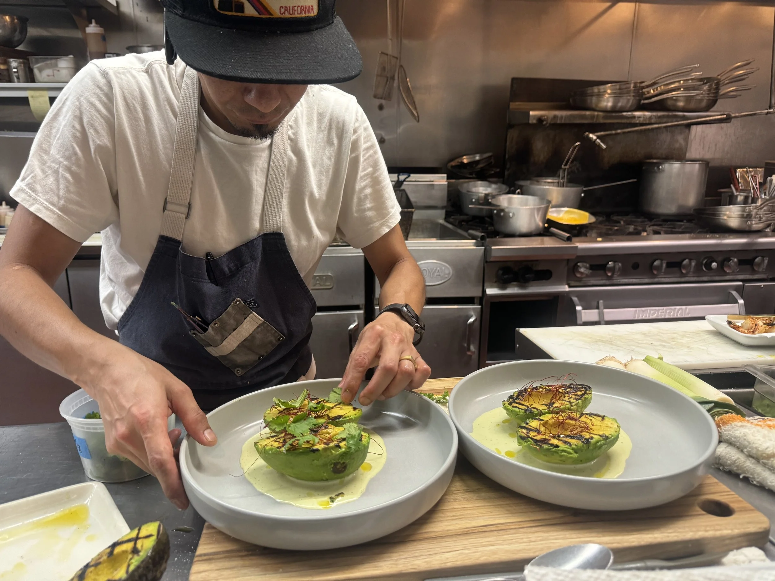 A chef garnishes two halved avocados with herbs and sauces on plates in a professional kitchen.