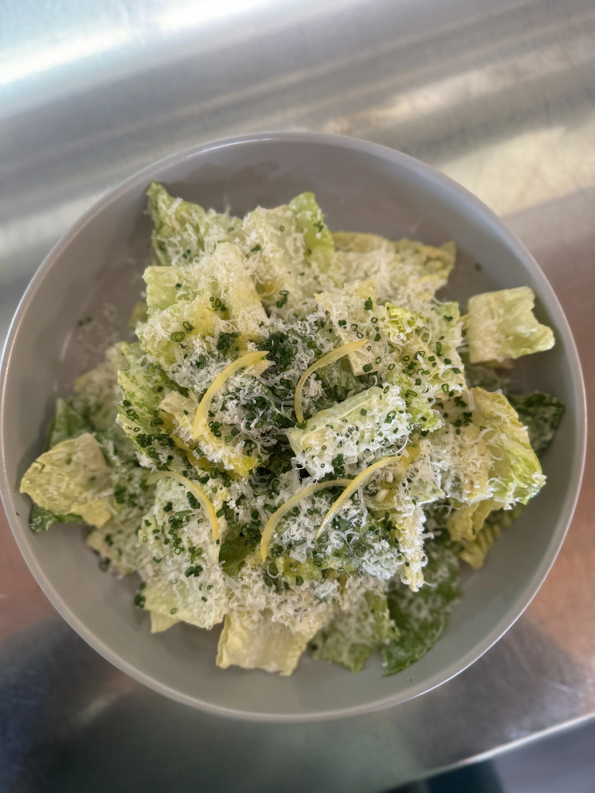 A bowl of chopped romaine lettuce, tossed with a creamy caesar dressing, topped with shredded cheese, lemon, and herbs.