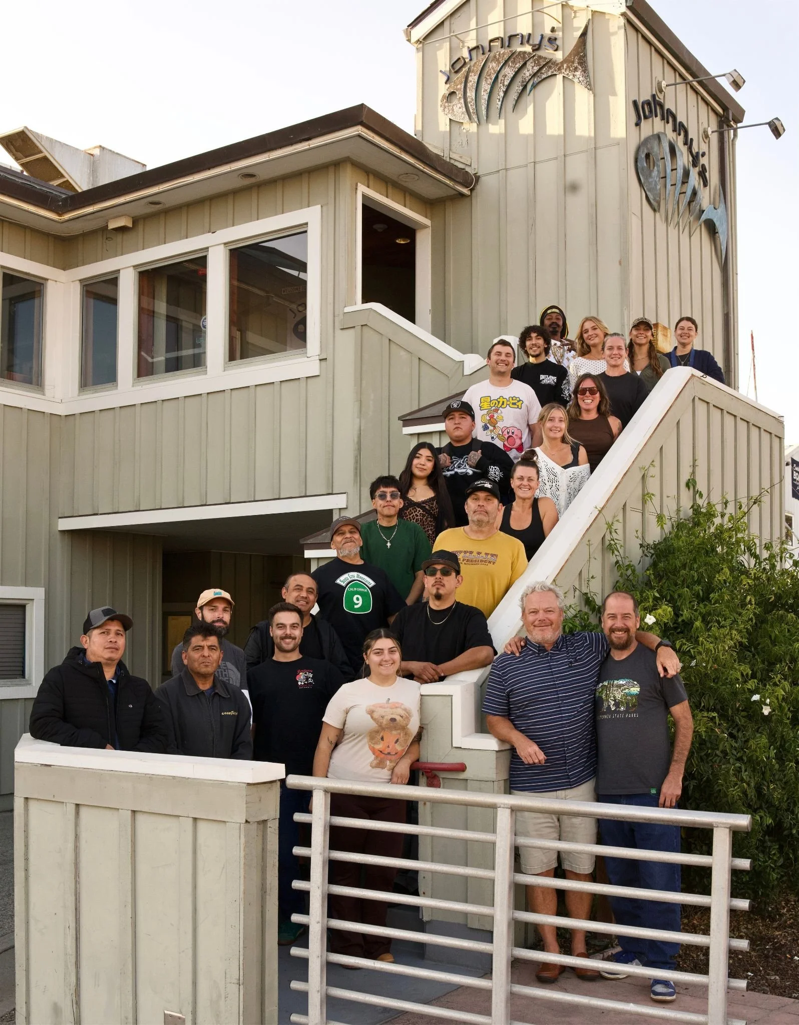 A group of people standing on the steps of a wooden building with a sign that reads 'Jonah's.' The group includes men and women of various ages, some smiling, some with serious expressions. One woman in the front is holding a stuffed animal. The building has large windows and a sign with a fish graphic. There are trees and plants around the building.
