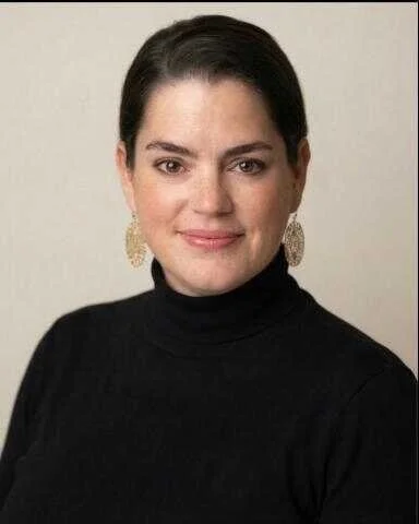 A woman with dark hair, wearing a black turtleneck and gold earrings, smiling at the camera against a plain background.
