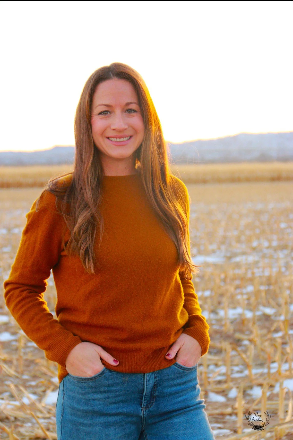 A woman with long brown hair smiling outdoors during sunset, wearing a brown sweater and blue jeans, standing in a field with some snow and dried plants, mountains in the background.
