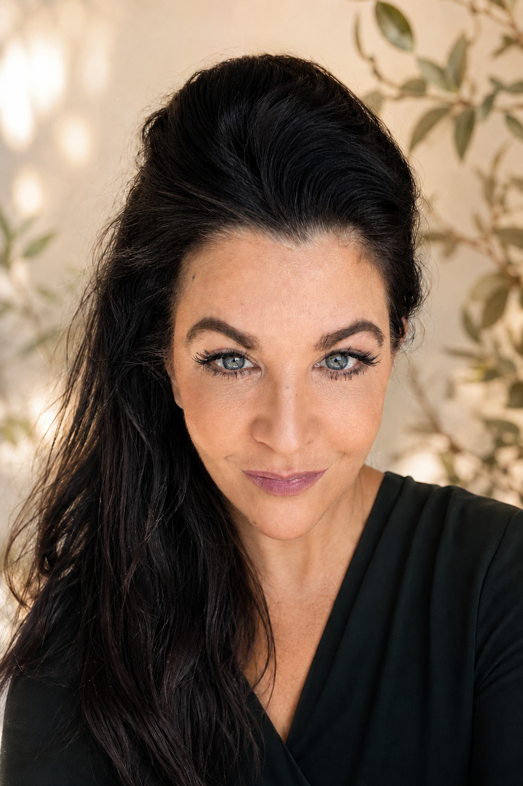 Un portrait d'une femme aux yeux bleus, cheveux noirs et longue, portant un haut noir, dans un décor avec des feuilles d'arbres en arrière-plan.