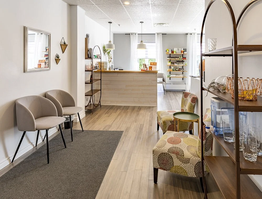 Salon d'attente dans un espace de bureau ou de clinique, avec des chaises beige, un tapis gris, des étagères en bois, et un comptoir de réception en bois clair. Éclairage lumineux naturel avec fenêtres et suspensions modernes. Espace Kinergie