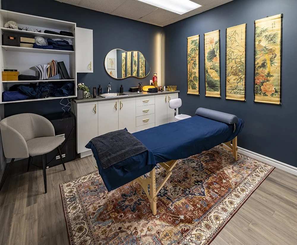 Une salle de traitement avec un lit de massage recouvert d'une drapée bleue, un tapis persan, un lavabo, un miroir, des étagères avec des serviettes et des articles, et des décorations murales aux motifs asiatiques. Espace Kinergie