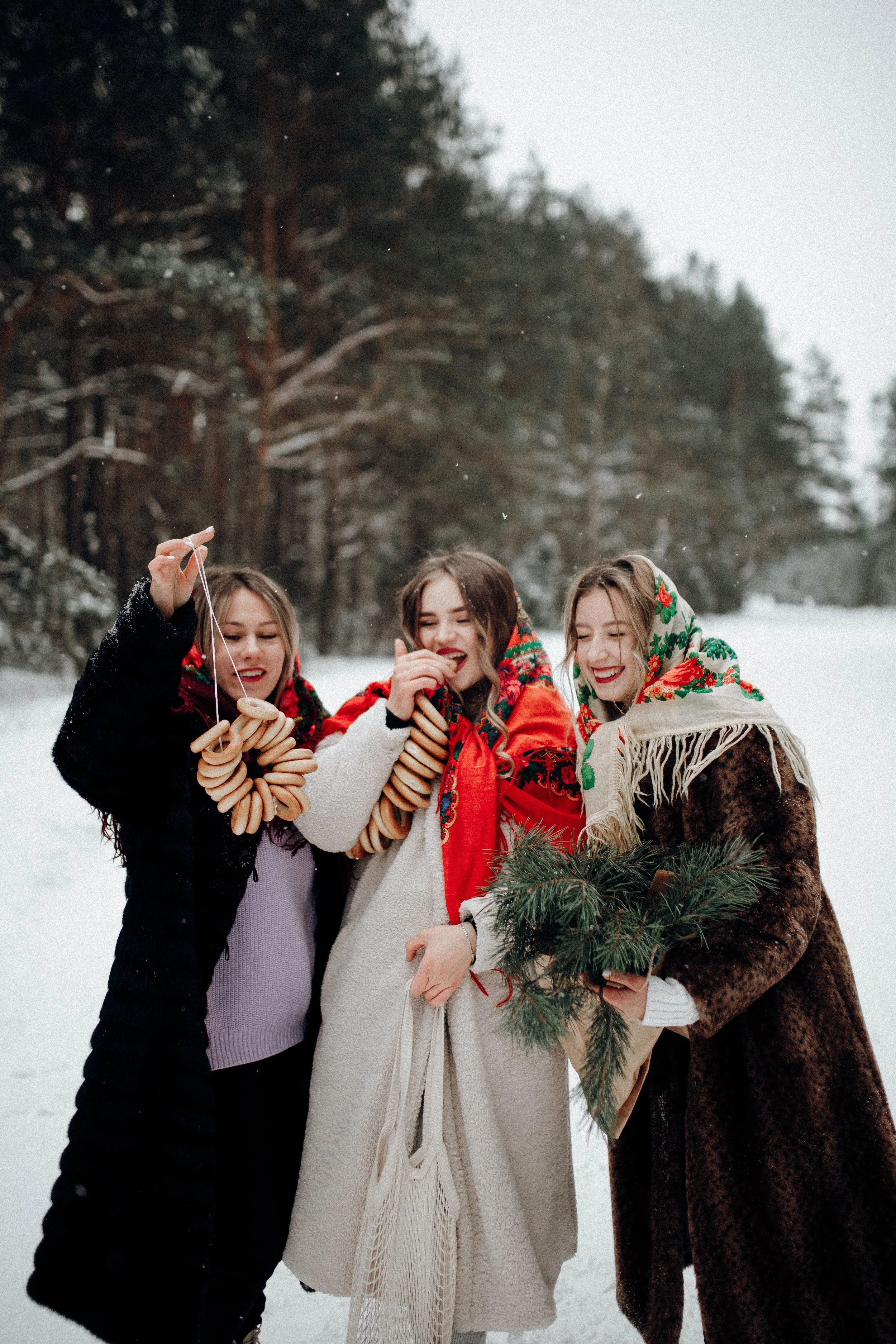 Three women in winter coats celebrate with pastries and pine branches, embracing the festive season.