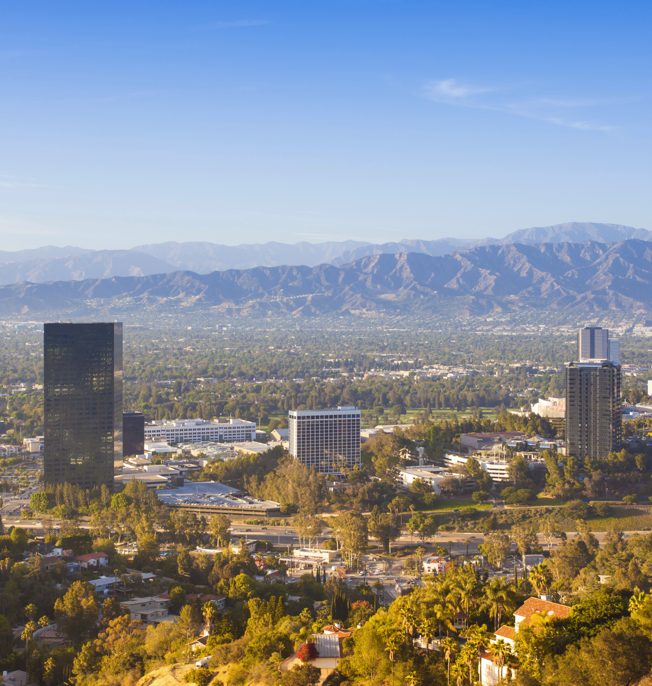 A cityscape with tall buildings and a highway, set against a background of mountain ranges and a clear blue sky.
