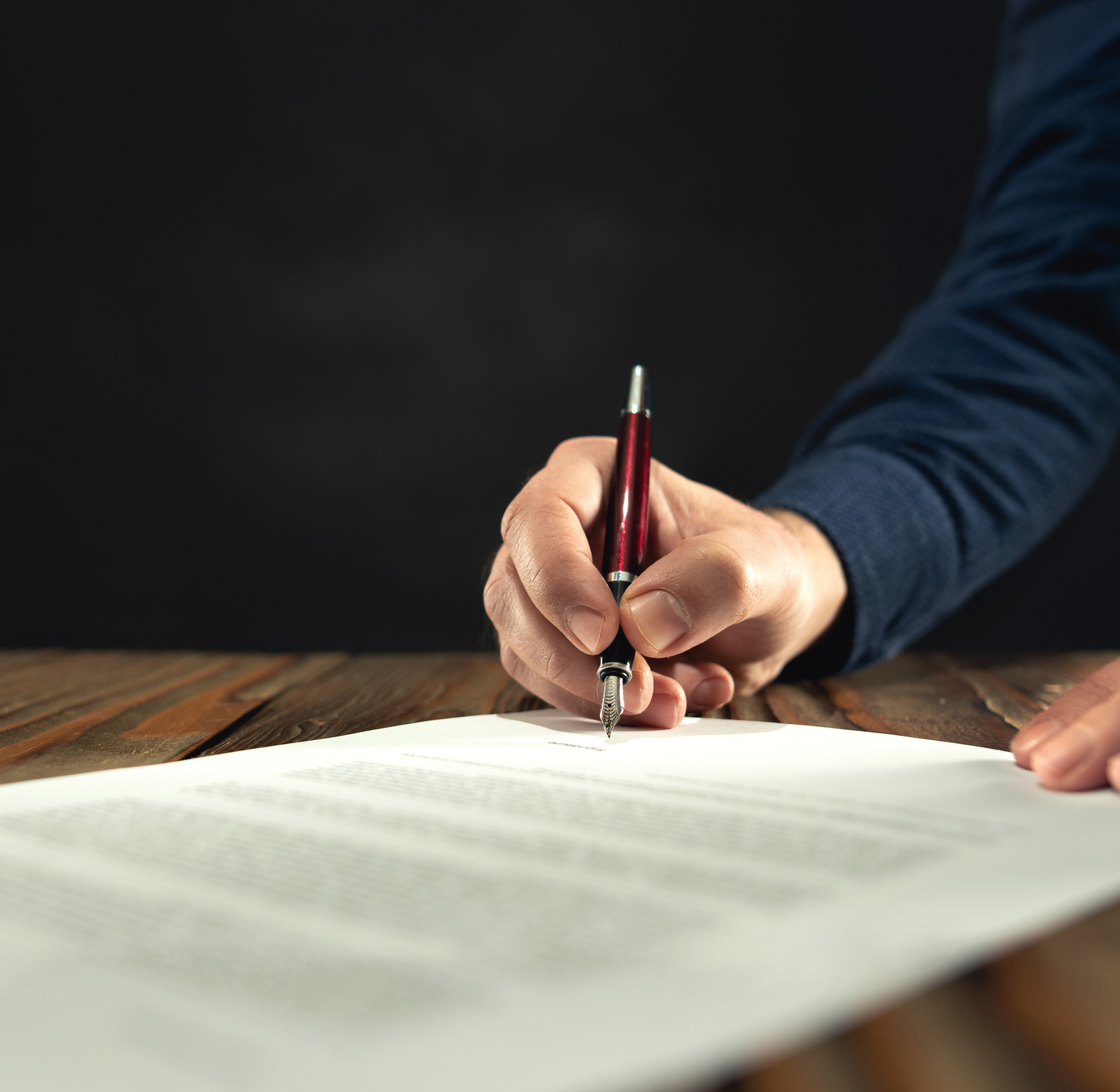 A person writing with a pen on paper. The person's hand is visible, and they are wearing a blue long-sleeve shirt. The background is dark and out of focus.