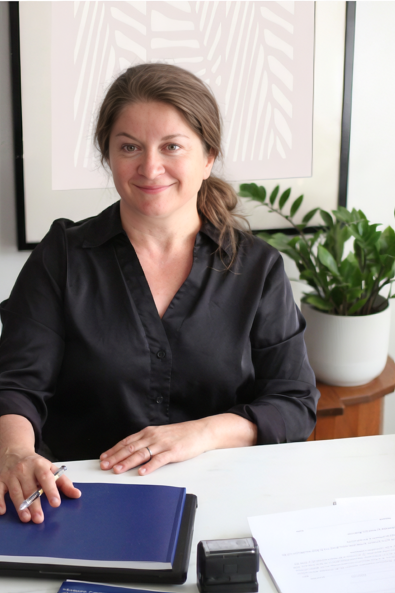 A woman with medium-length brown hair smiling, wearing a black blouse, sitting at a white table with a blue folder, papers, a pen, and a black stamp. Behind her, there is framed abstract art and a potted plant.