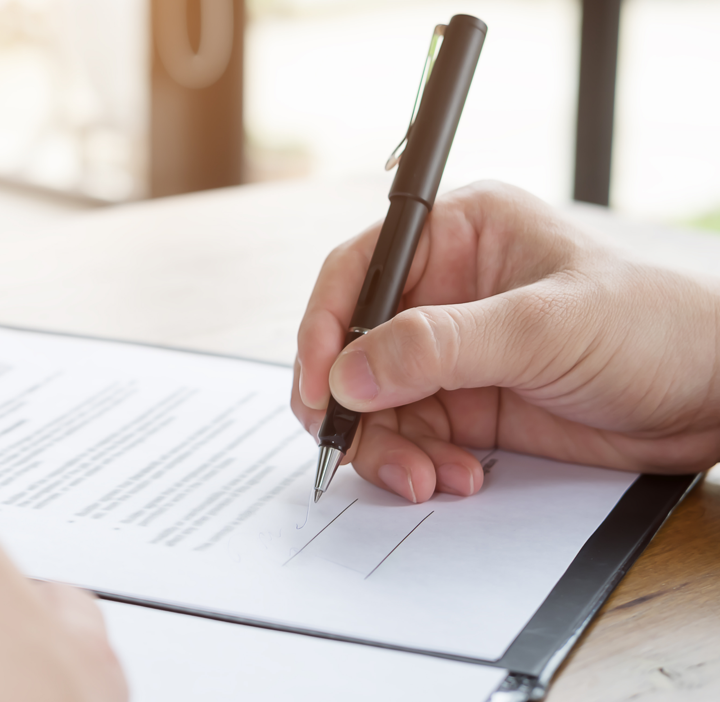 Close-up of a person's hand writing on lined paper with a black pen, with sunlight streaming through a window in the background.