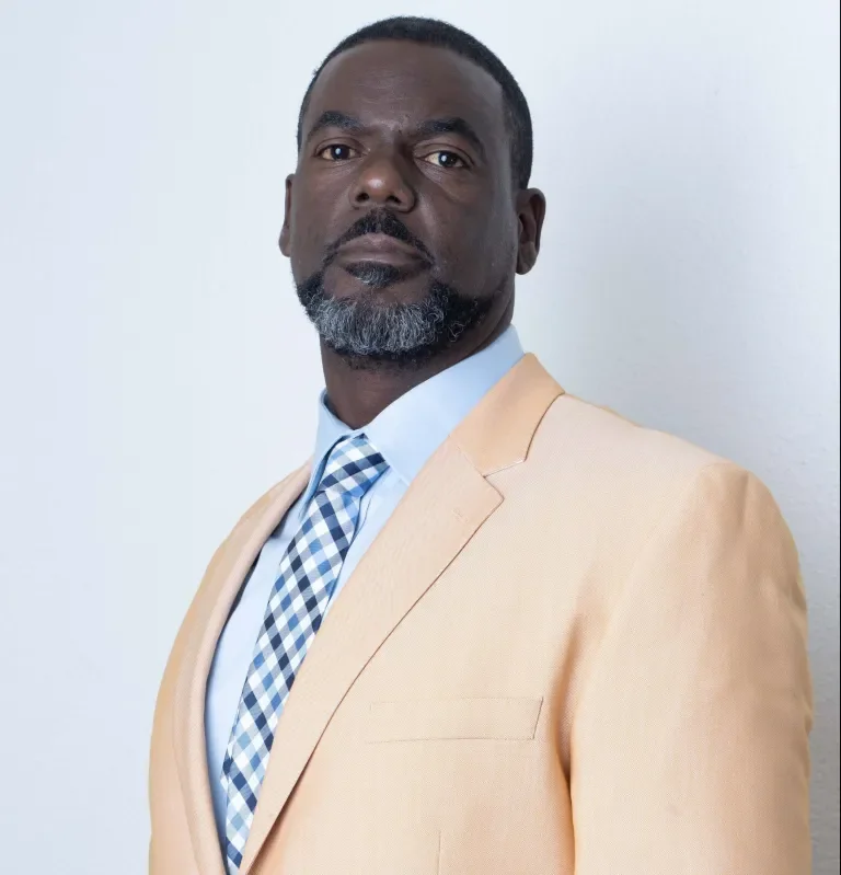 A confident man wearing a beige suit jacket, light blue dress shirt, and checkered blue and white tie, standing against a plain white background.