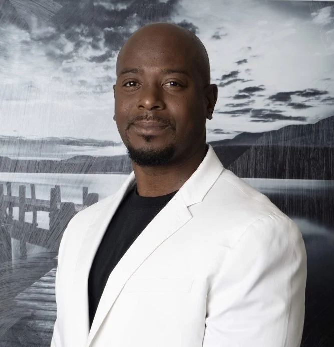 A confident man with a bald head and goatee wearing a white blazer over a black shirt, standing in front of a scenic backdrop of a dock and mountains under a cloudy sky.