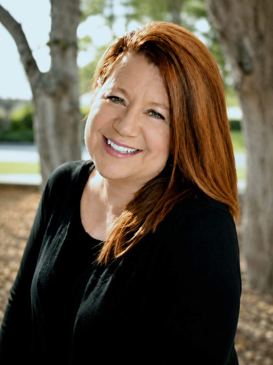 A confident smiling woman with red hair outdoors near trees and water.