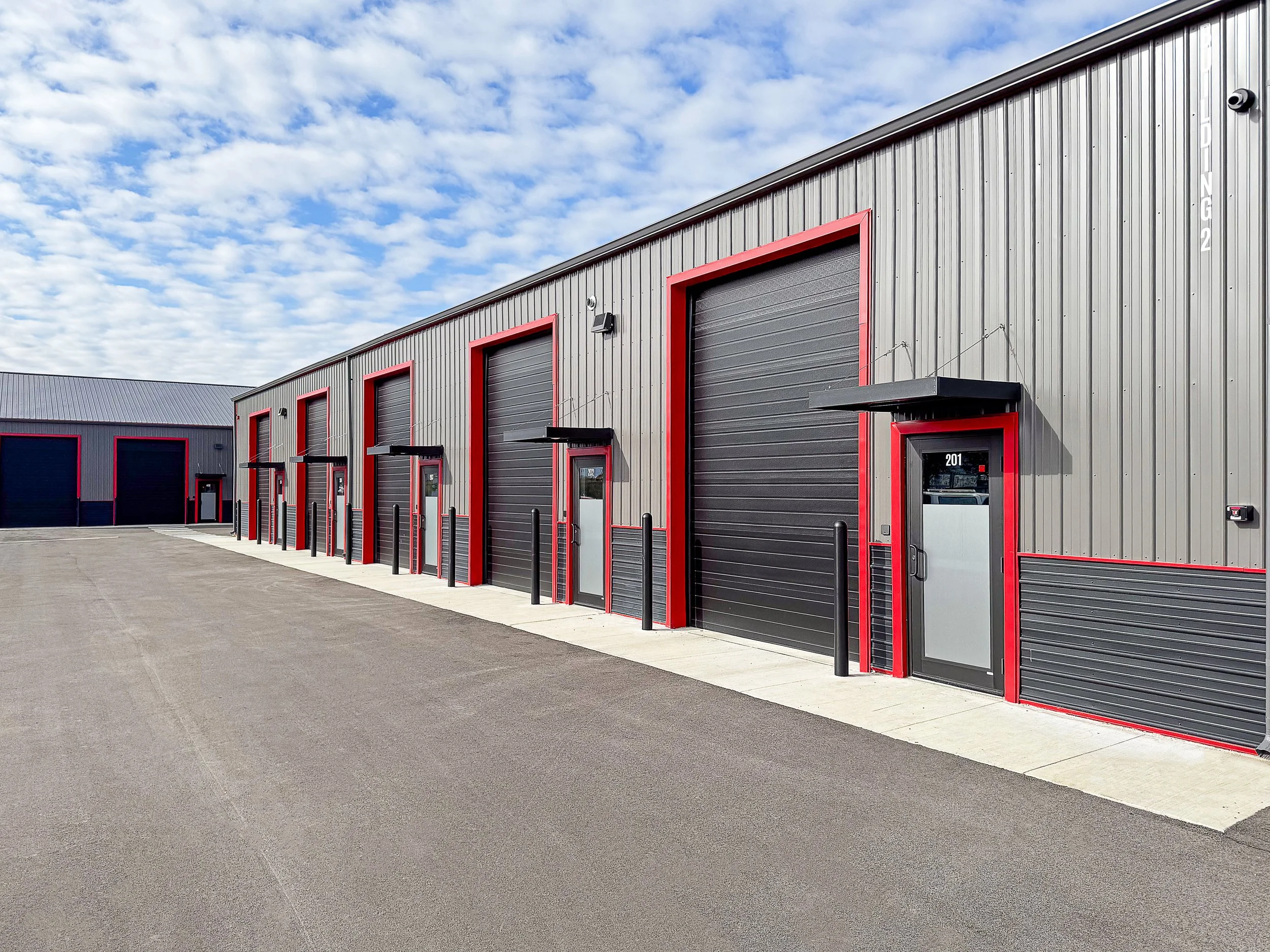 A modern commercial self-storage facility with multiple gray metal units bordered with red trim, black roller doors, and a clear sky with scattered clouds overhead.