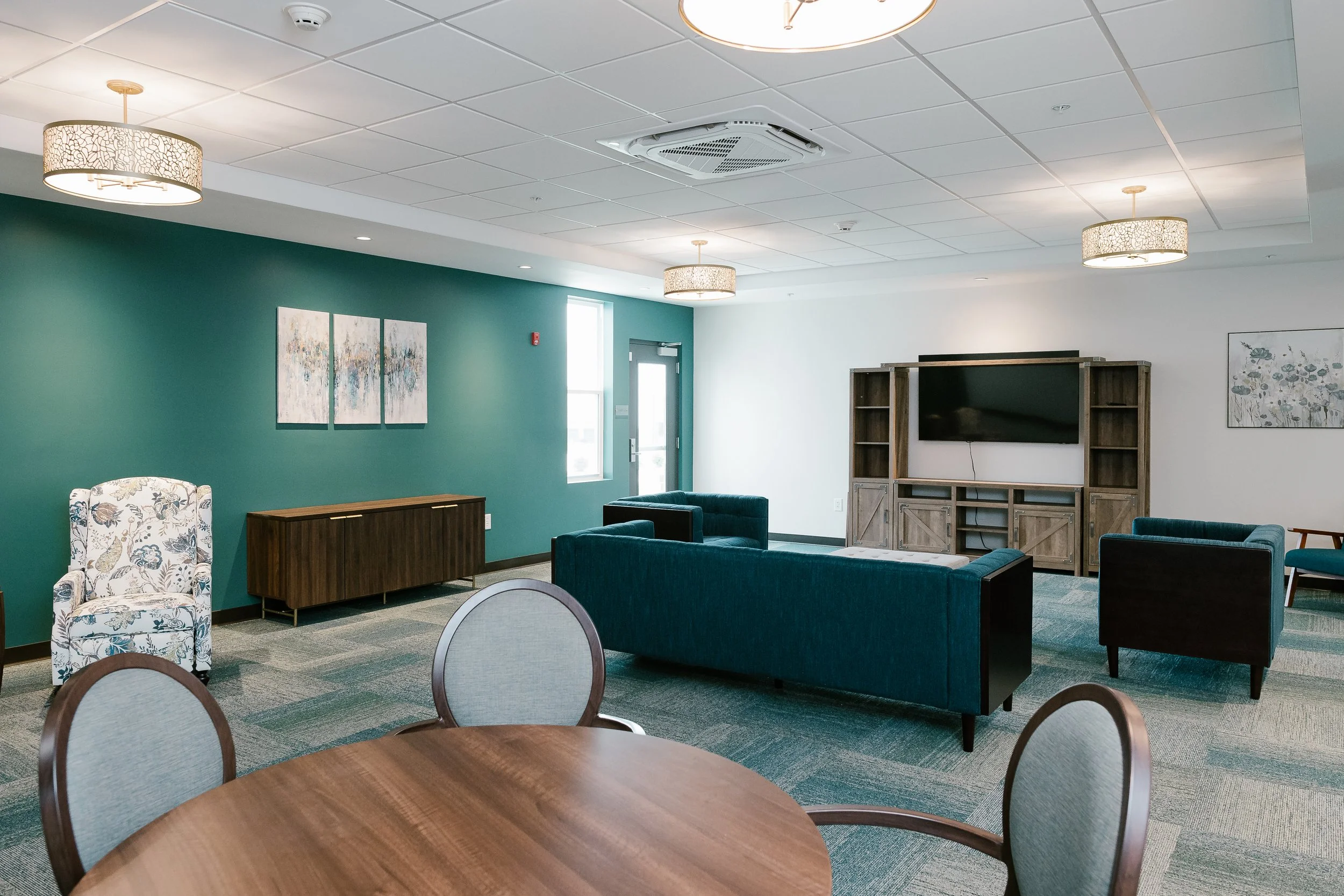 A modern lounge room with blue walls, circular ceiling lights, a floral armchair, a wooden sideboard, several blue sofas, a wall-mounted flat-screen TV, and framed floral artwork.