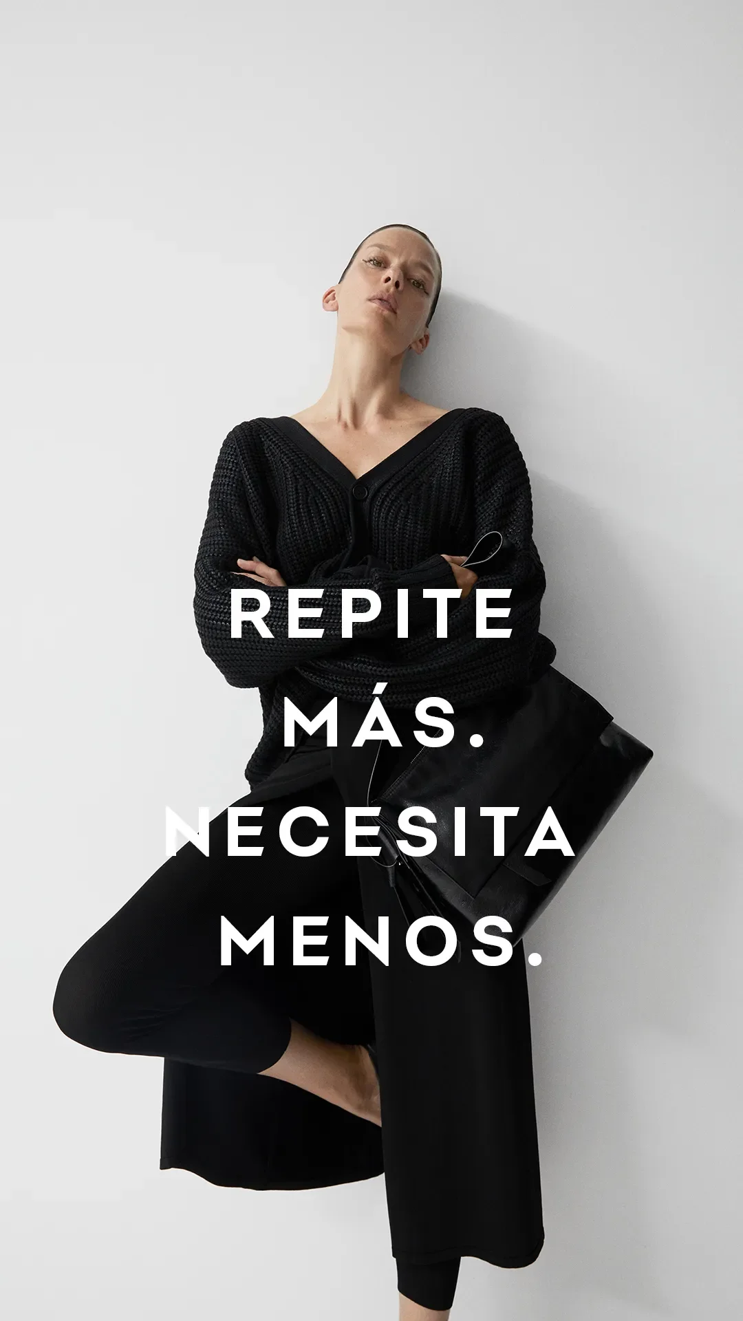 Una mujer con expresión seria viste ropa negra y sostiene una bolsa negra, con un fondo gris claro. Texto en español: "Repite más. Necesita menos."
