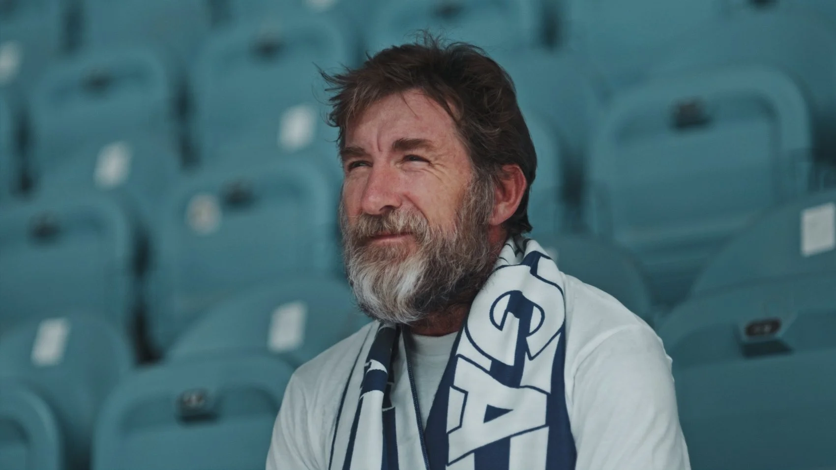 Hombre con barba y cabello castaño, sentado en una grada de estadio, vistiendo una camiseta blanca y una bufanda azul y blanca.