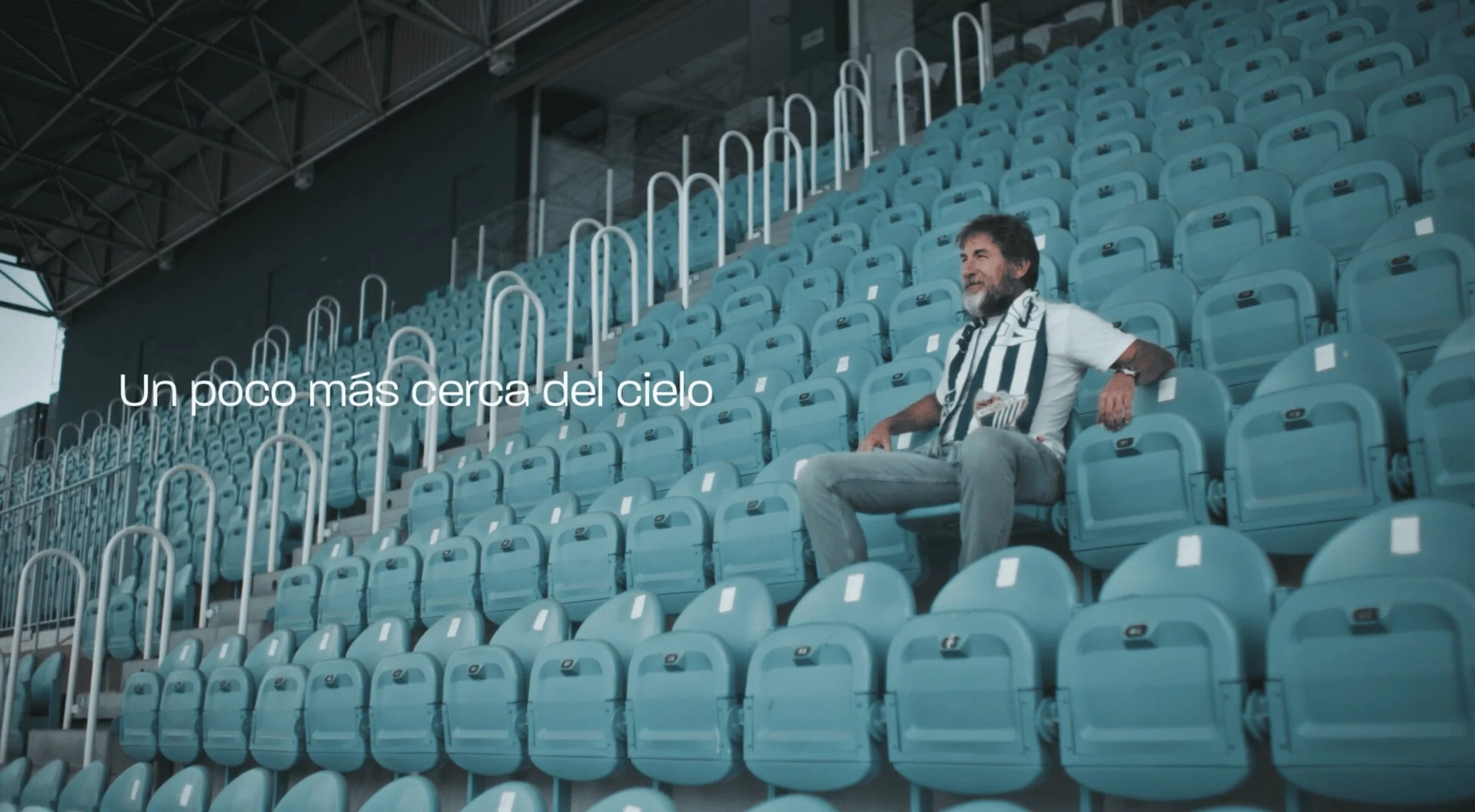 Hombre sentado en asientos vacíos de un estadio, con la frase en español 'Un poco más cercano del cielo'