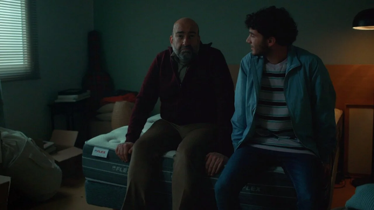 Dos hombres sentados en una cama en un cuarto oscuro, uno parece mayor y el otro joven, hablando entre sí.