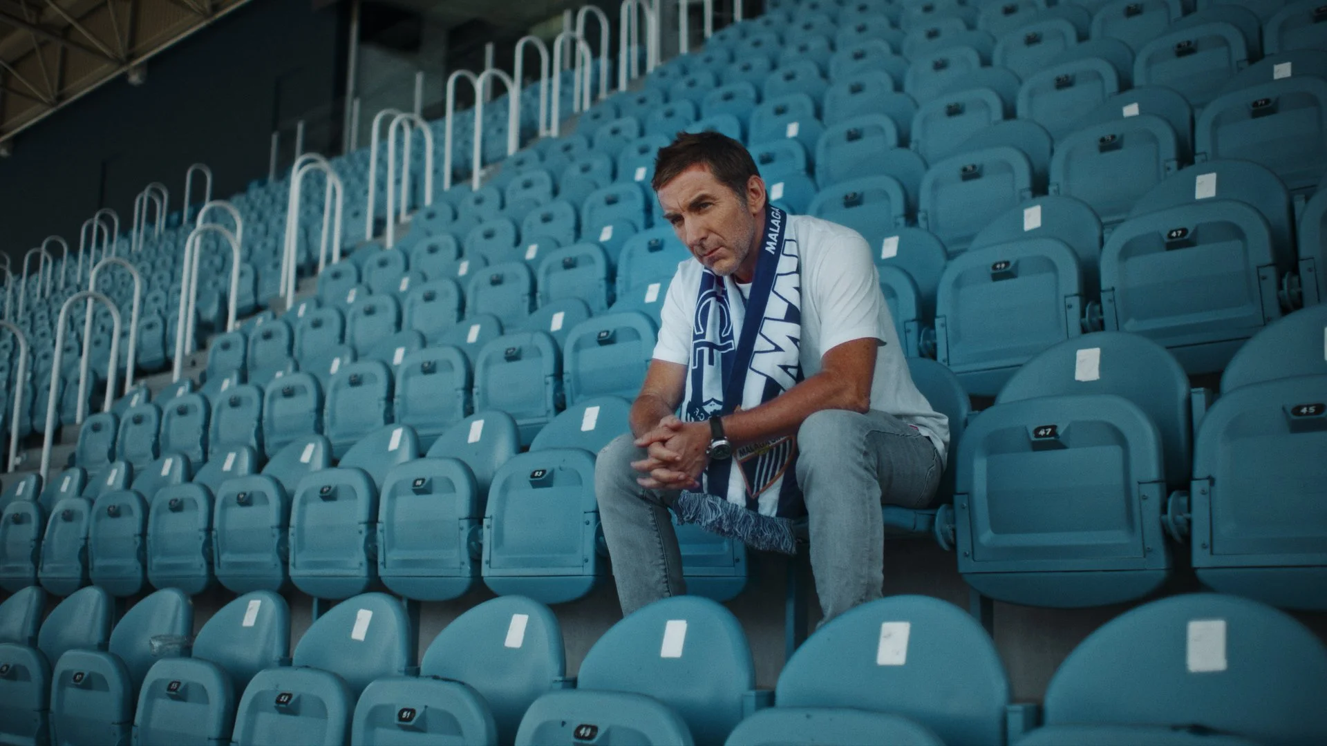 Hombre sentado en asientos de un estadio vacío, usando una bufanda azul y blanca, vestimenta casual, con expresión pensativa.