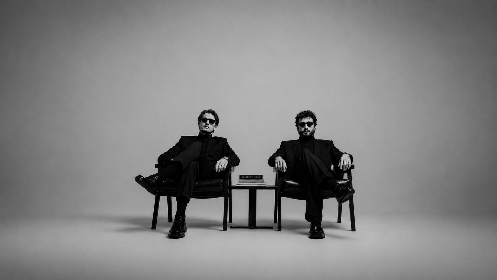 Dos hombres en gafas oscuras sentados en sillas negras, con un libro sobre la mesa entre ellos, en un fondo gris liso.